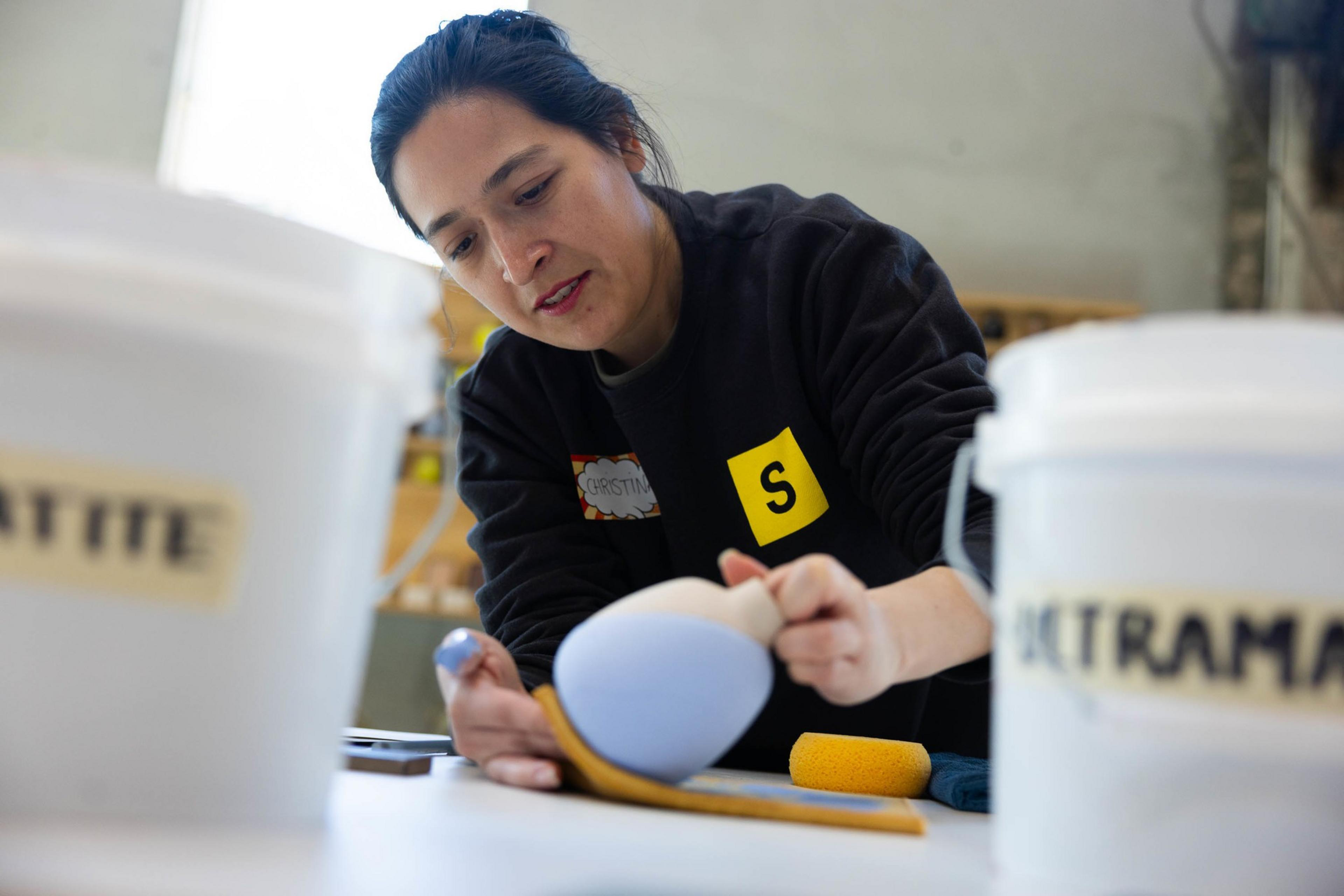 A woman is attentively painting a blue bud vase on a workbench with containers labeled "WATER" and "ULTRAMARINE" nearby.