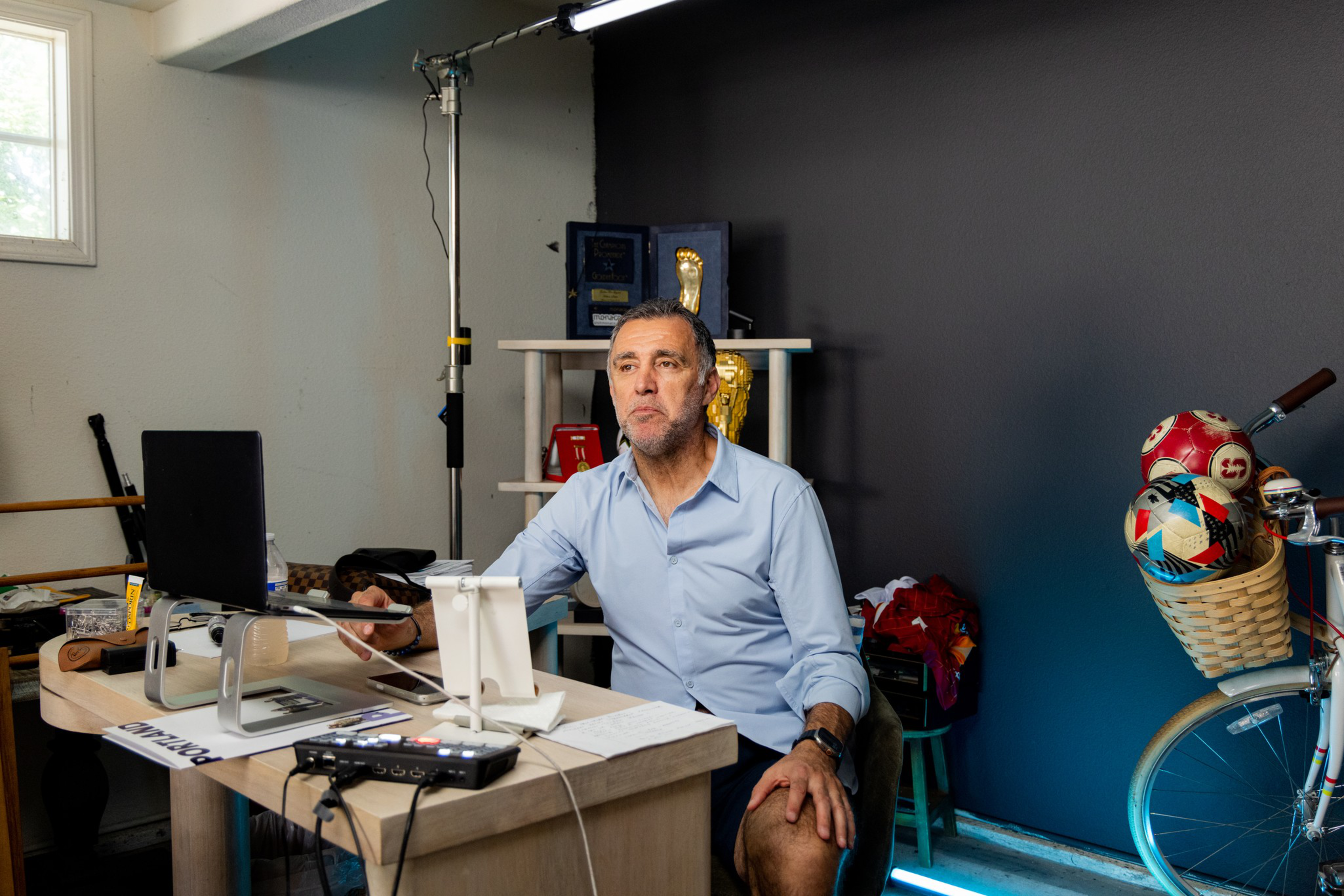 A man in a light blue shirt sits at a desk with a laptop and audio equipment, a bicycle with soccer balls in its basket is beside him.