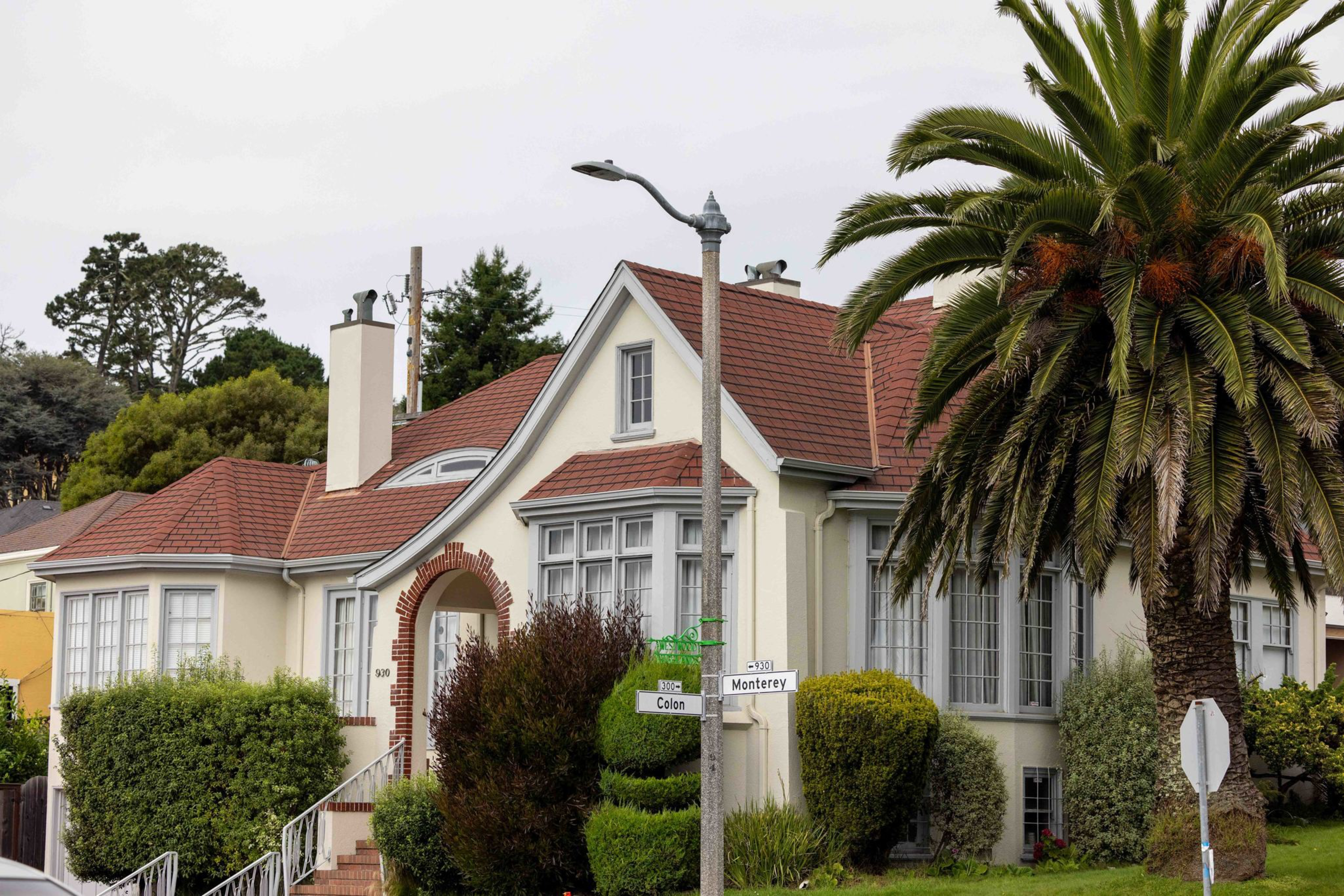A beige house with a red-tiled roof is surrounded by trimmed bushes and a tall palm tree, located at the intersection of Colon and Monterey streets.