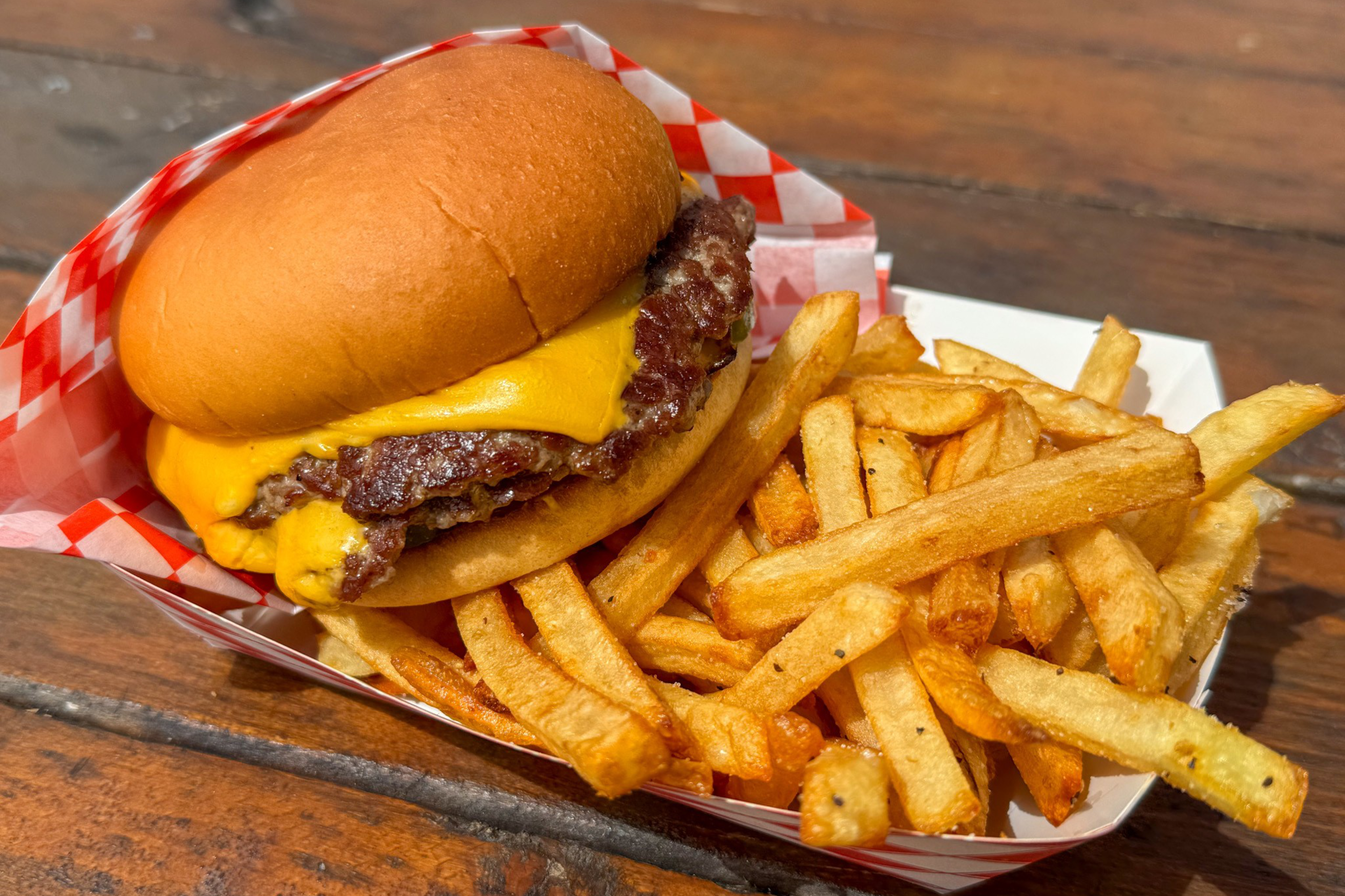 A cheeseburger with fries in a checkered basket, on a wooden surface.