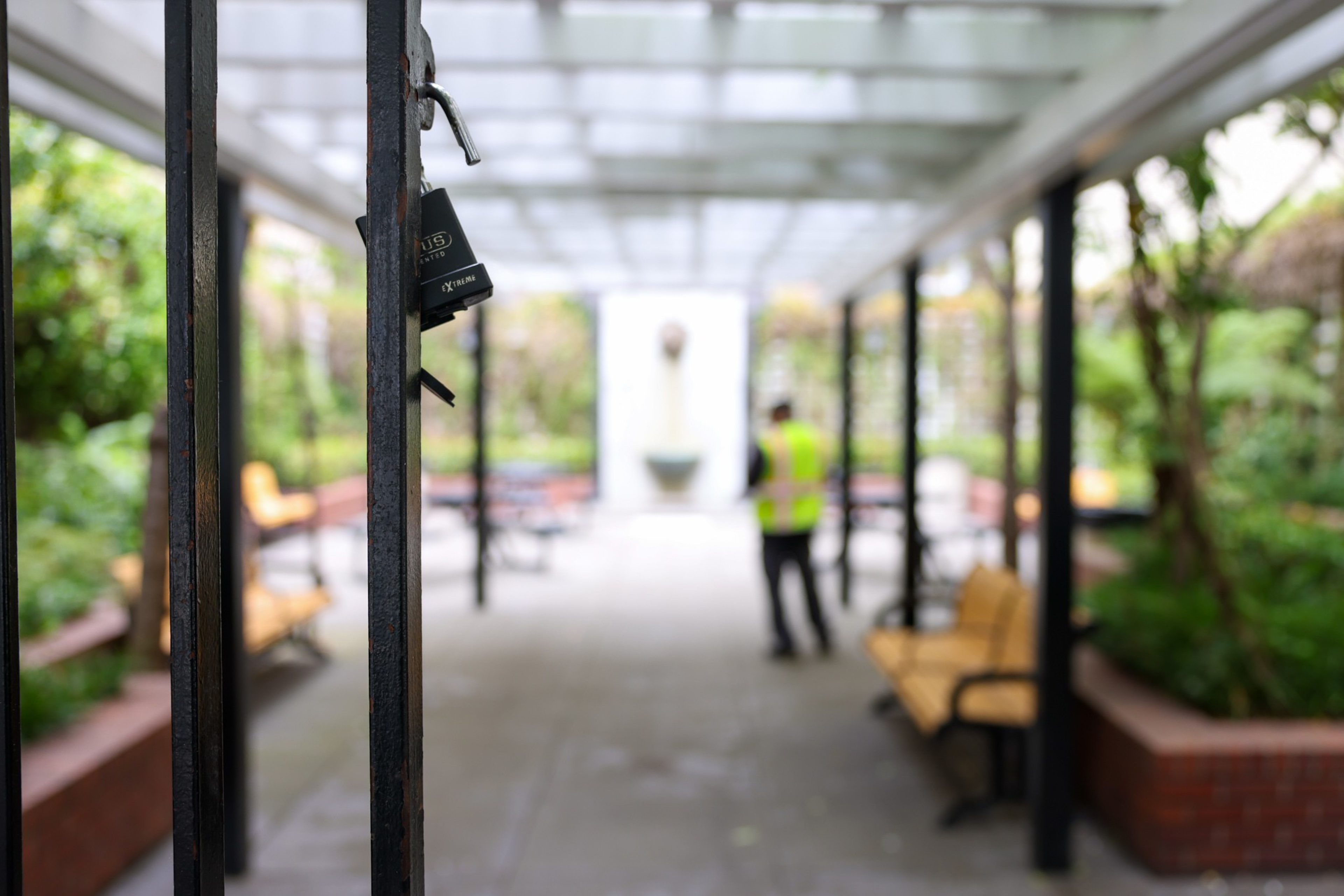 A close-up of a black metal gate with a lock in front of a covered outdoor walkway, with blurred benches, plants, and a person in a neon vest in the background.