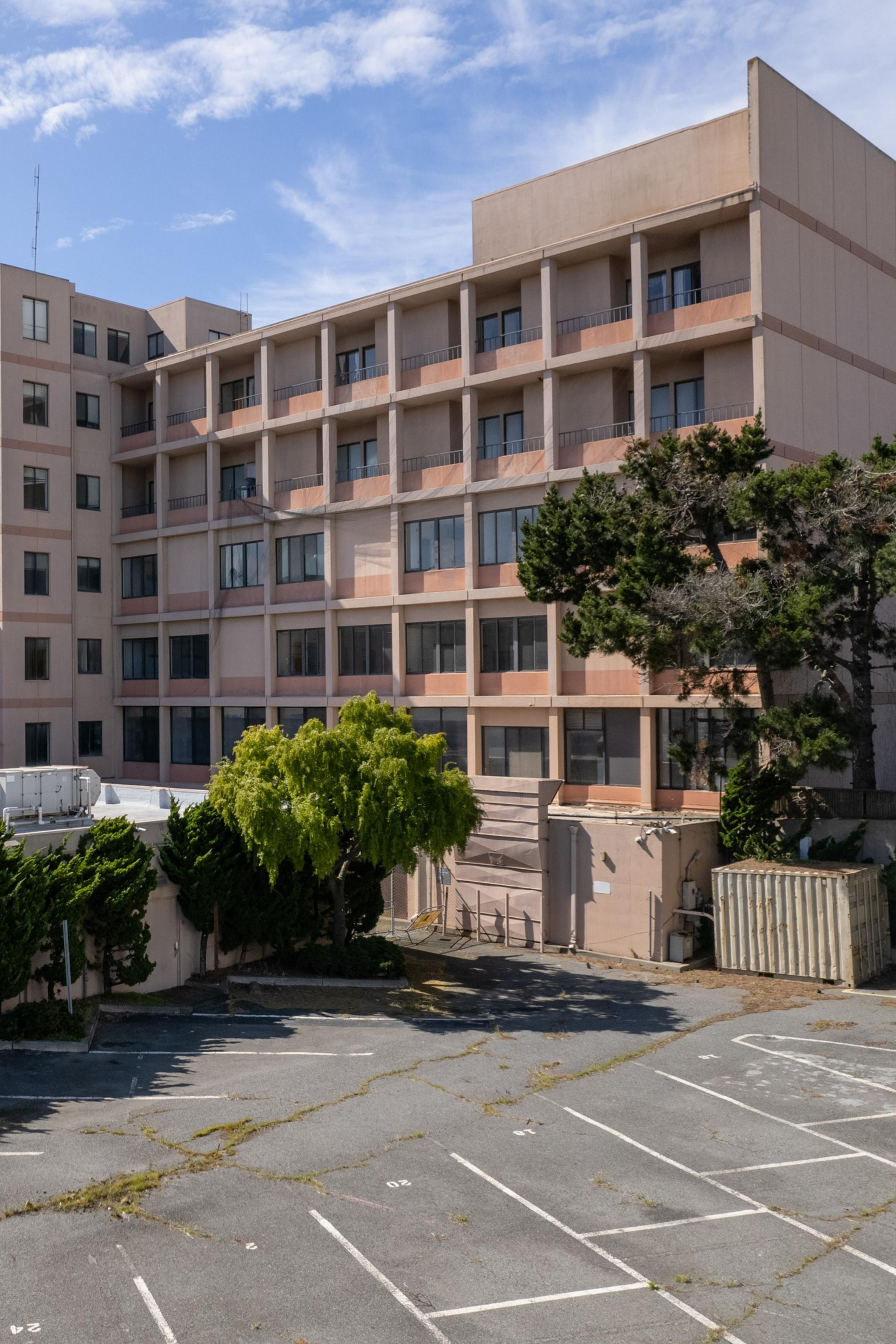 A five-story beige building with balconies, a parking lot in front with no cars, and a few green trees under a blue sky.