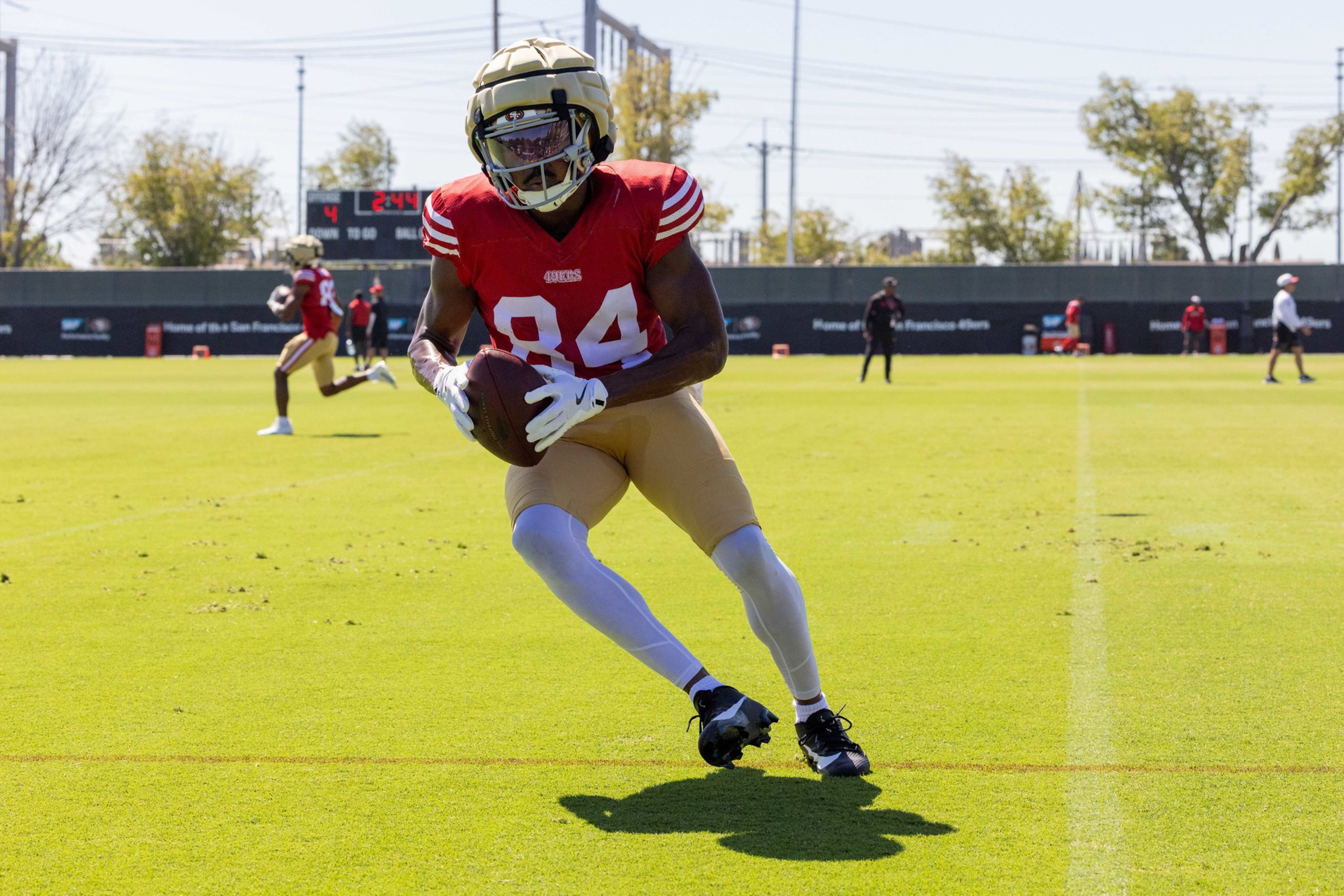 A football player in a red jersey and gold pants runs on a field, holding a football, wearing a helmet and white gloves during practice.