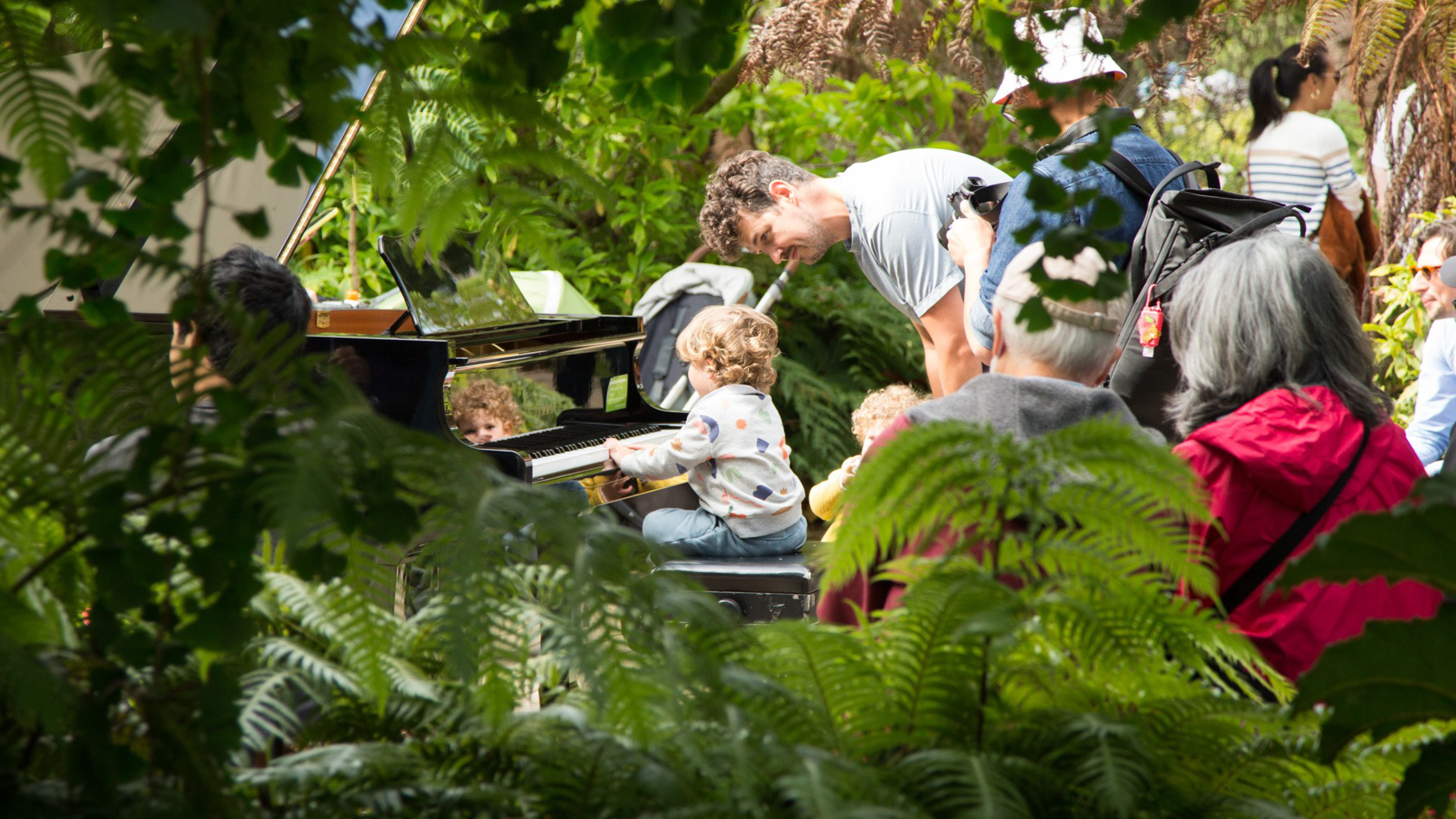 A young child plays a piano outdoors surrounded by lush green plants while adults watch, including a man bending down to assist the child.
