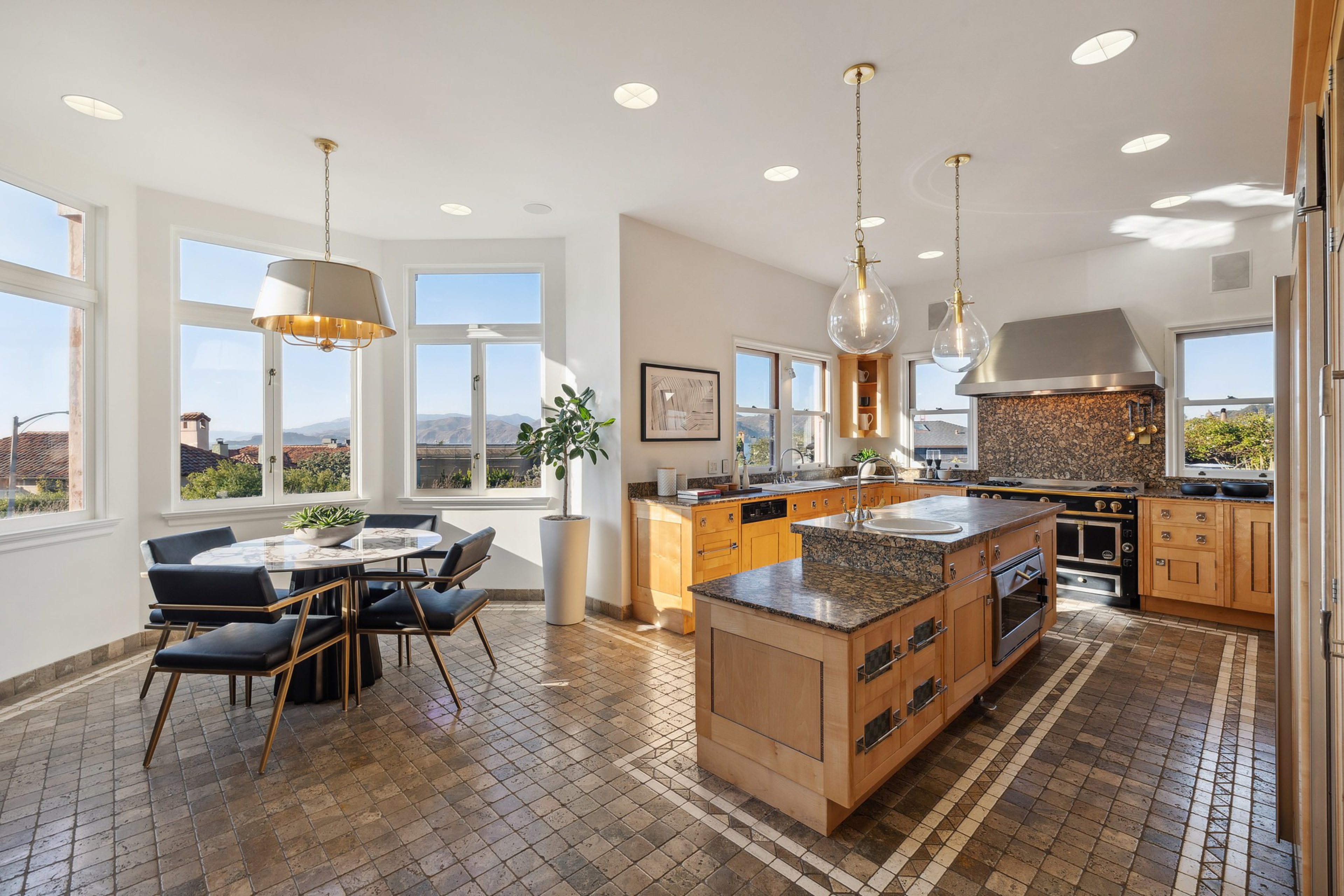 A spacious kitchen with tiled floors, wooden cabinets, a granite island, stainless steel appliances, and a dining table surrounded by black chairs by large windows.