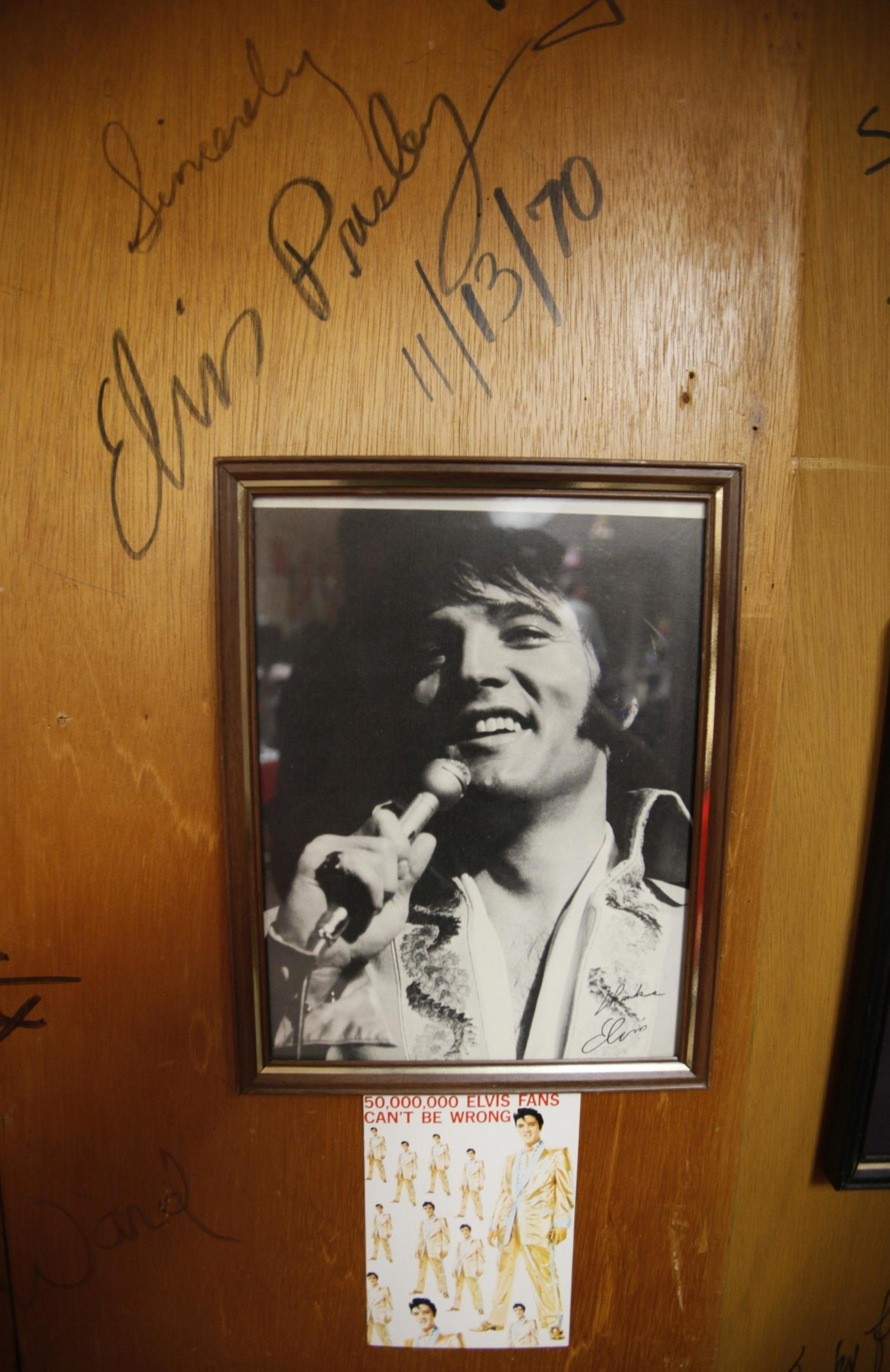 A framed black-and-white photo of a smiling man holding a microphone, signatures, and an &quot;Elvis fans&quot; sticker on a wooden background. Elvis's signature appears on the wall above the photo.