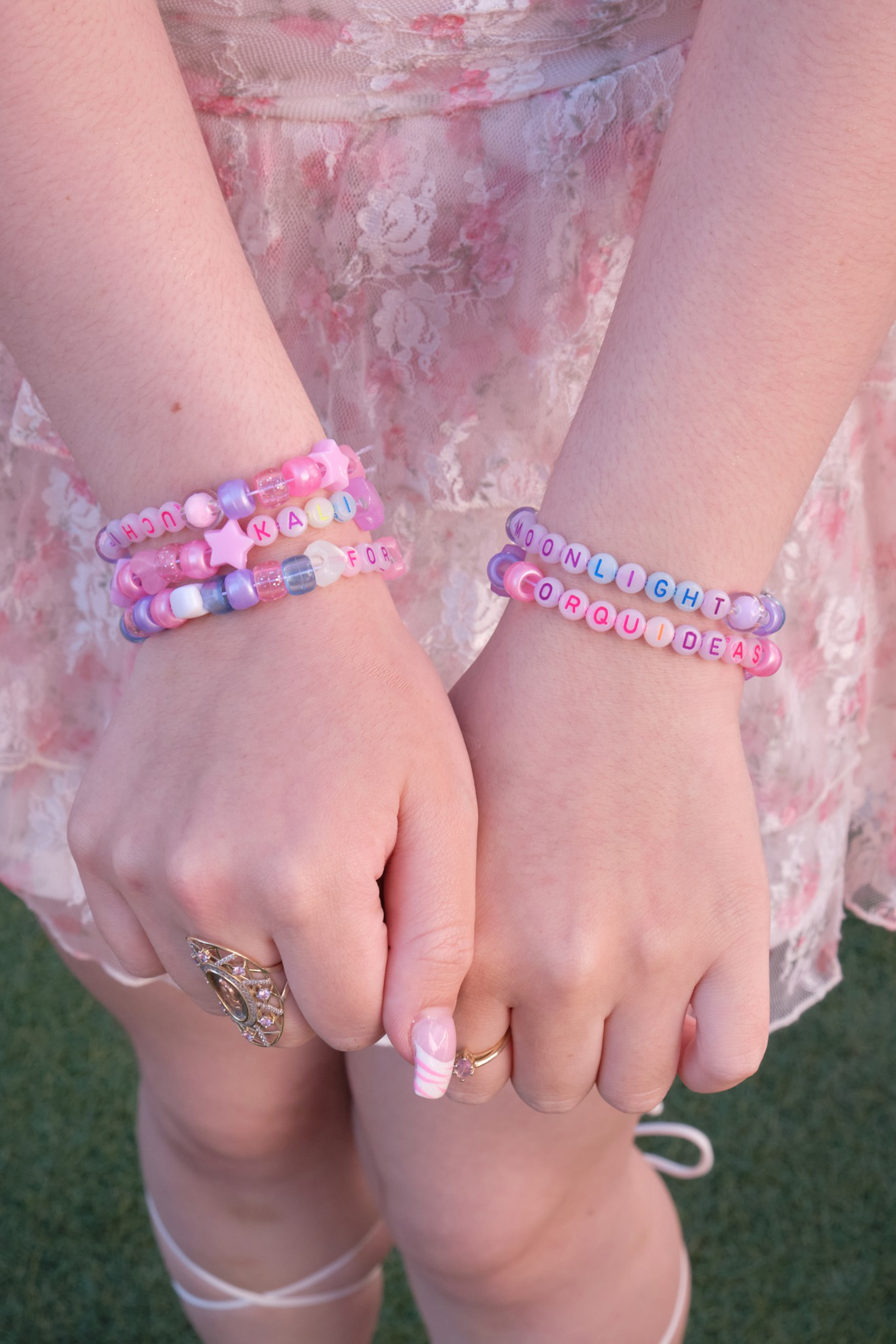 Two hands with pink-themed beaded letter bracelets that spell out words like "MOONLIGHT" and "CORQUIDEAS" rest over a pink floral lace dress.