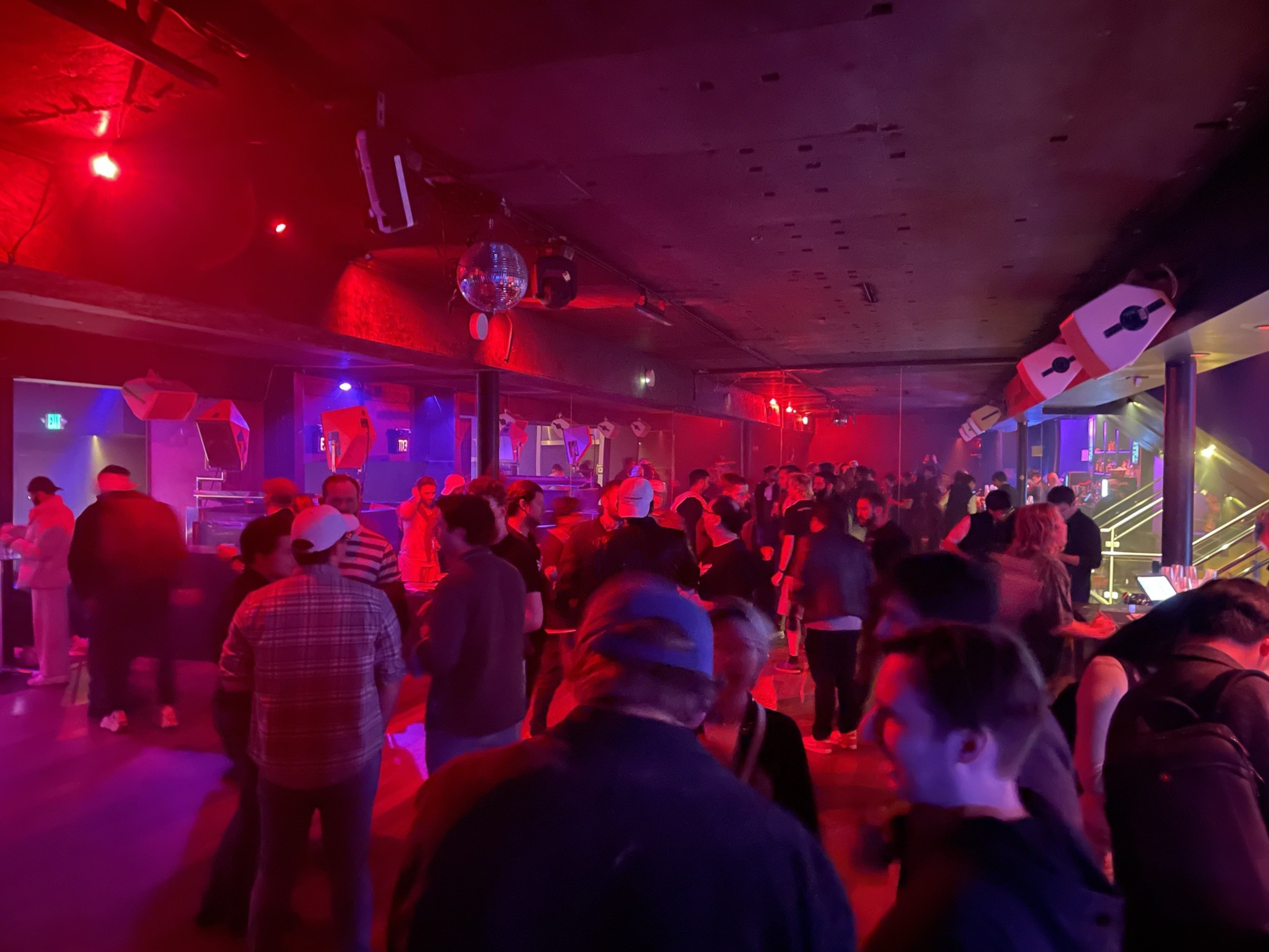A large crowd of more than 100 people are seen standing with their backs turned and bathed in red and blue lights near a bar inside a nightclub with large speakers hanging overhead.
