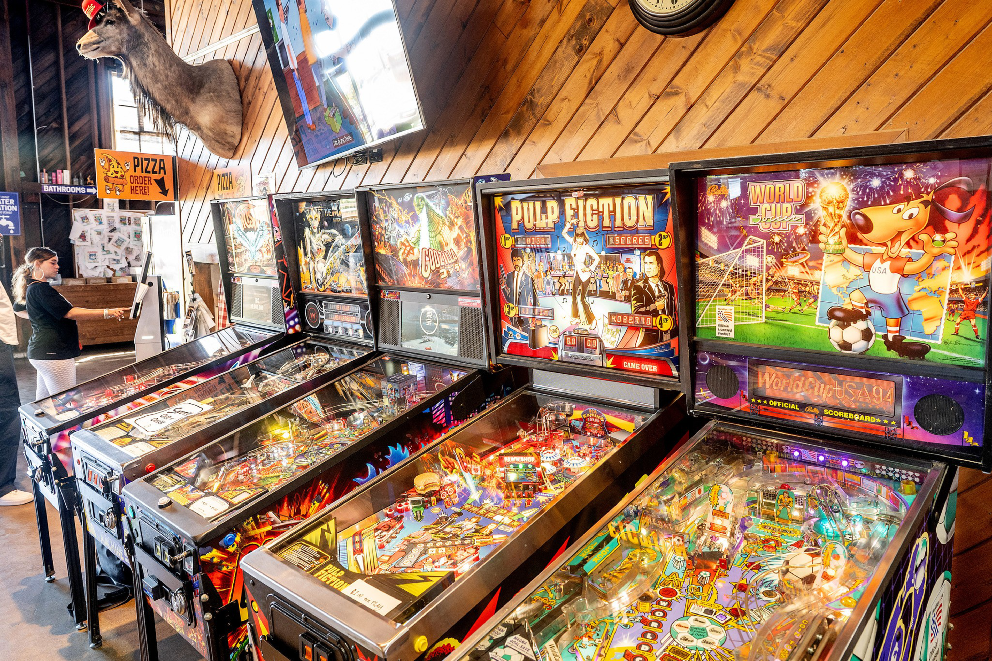 A row of colorful pinball machines lines a wooden wall with a mounted deer head above and a woman playing in the background.