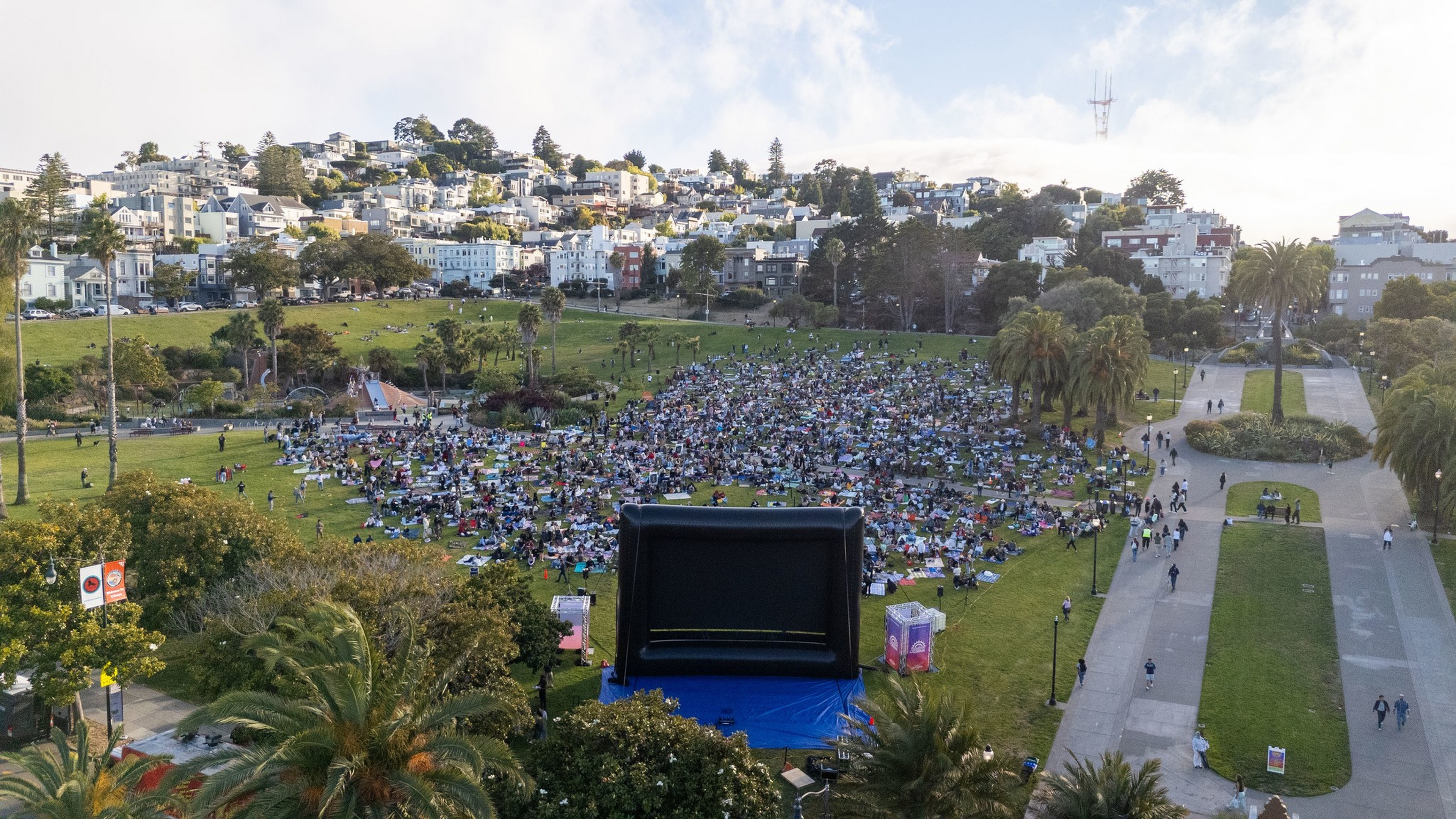 A large crowd gathers on a grassy hill for an outdoor movie screening, with an inflatable screen and city buildings visible in the background.
