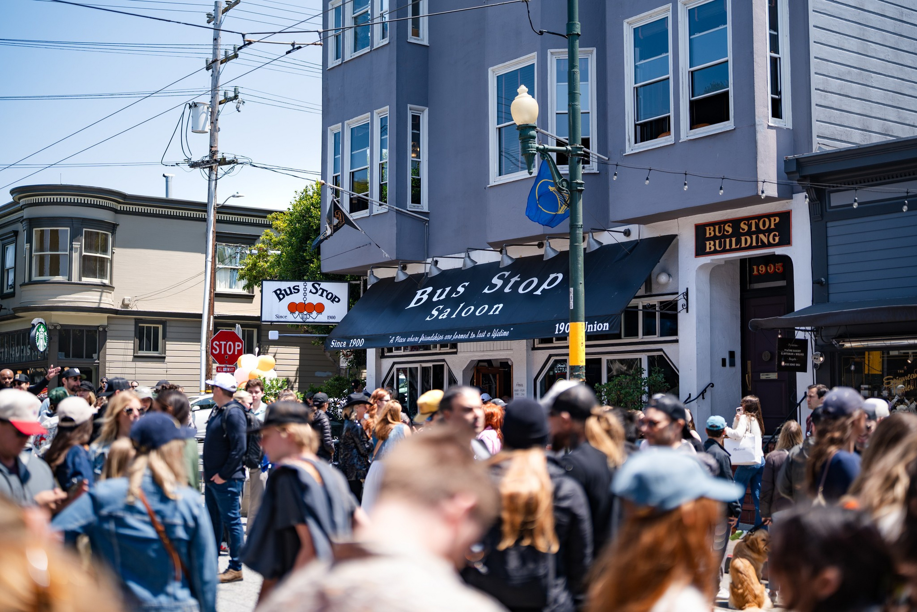 A large crowd gathers outside the Bus Stop Saloon on a sunny day at a busy street corner with power lines overhead.