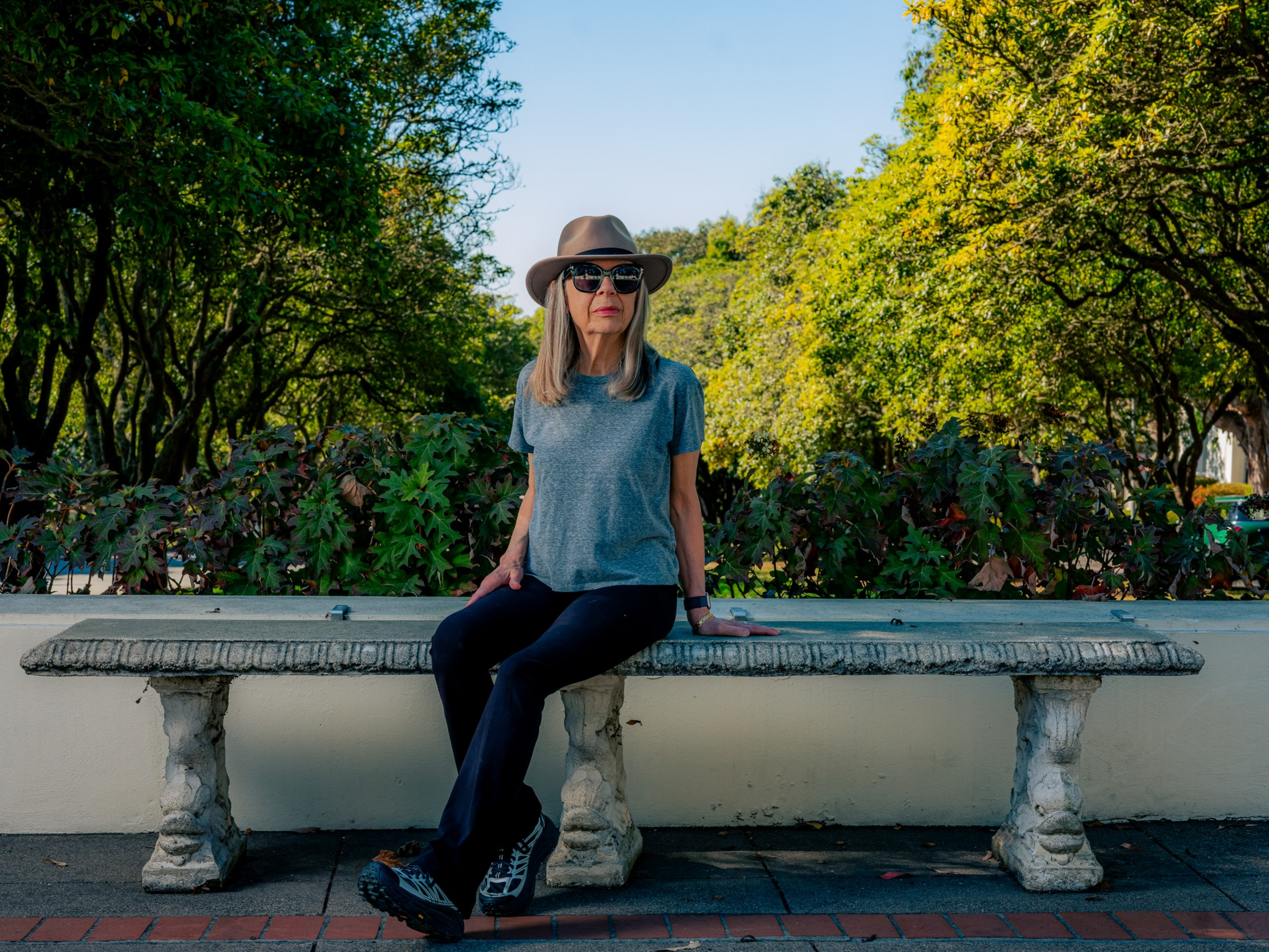 A woman wearing a gray shirt, black pants, sunglasses, and a hat sits on a stone bench surrounded by green foliage and trees.