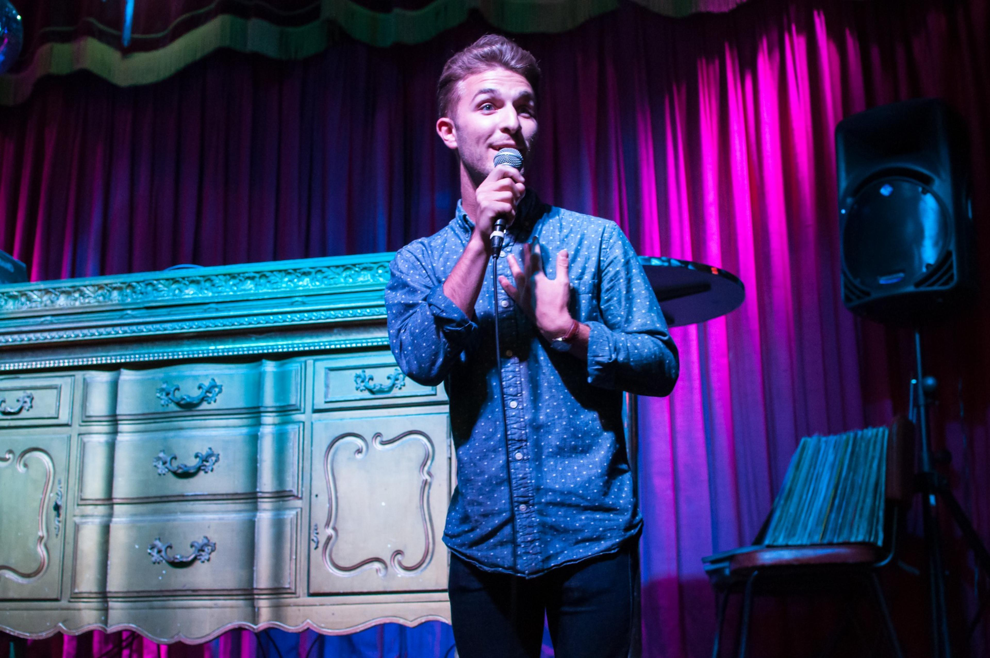 A man with a mic stands in front of a dresser on a stage bathed in purple light.