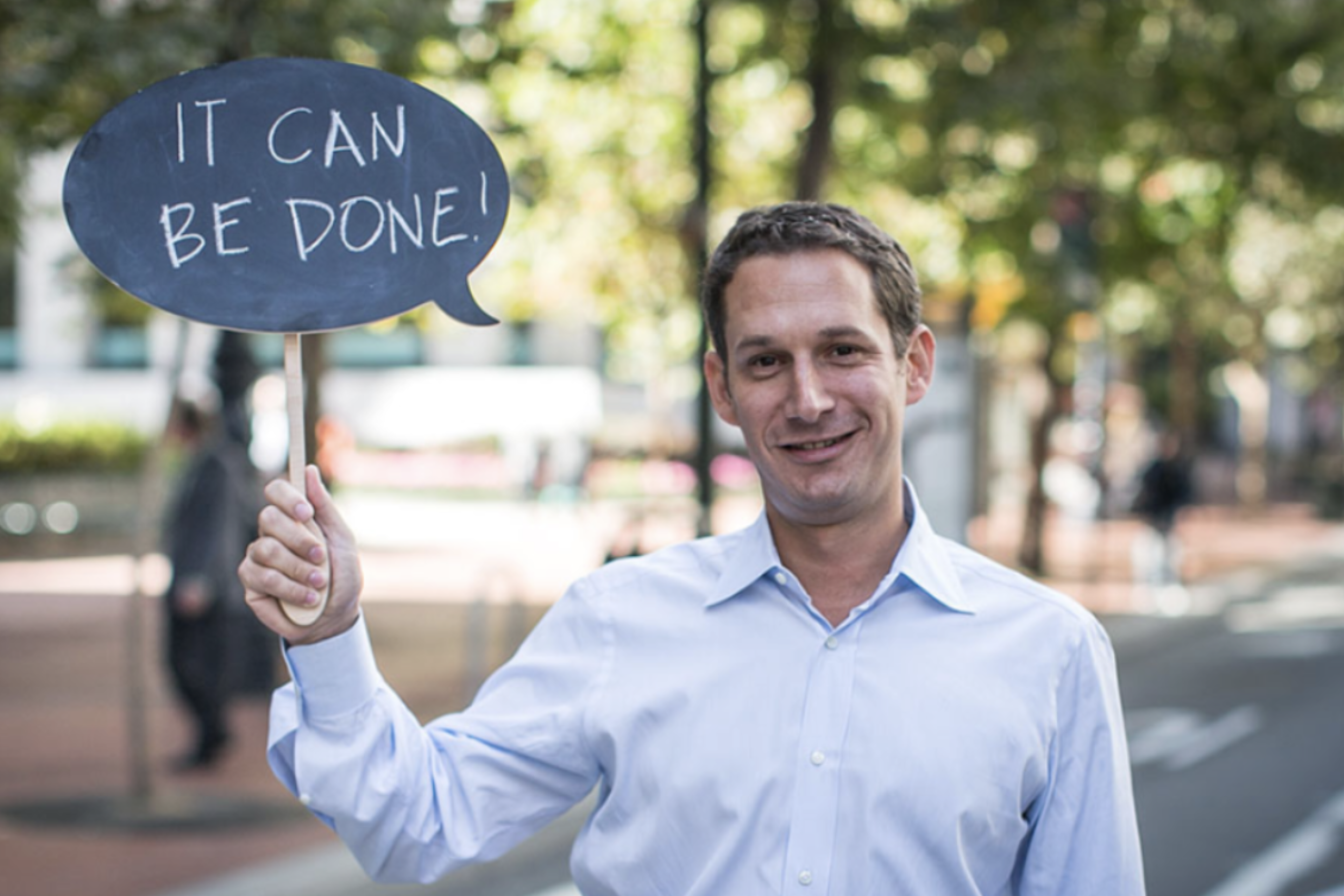A man in a light blue shirt smiles and holds a speech bubble sign that reads "IT CAN BE DONE!" with a blurred outdoor background.