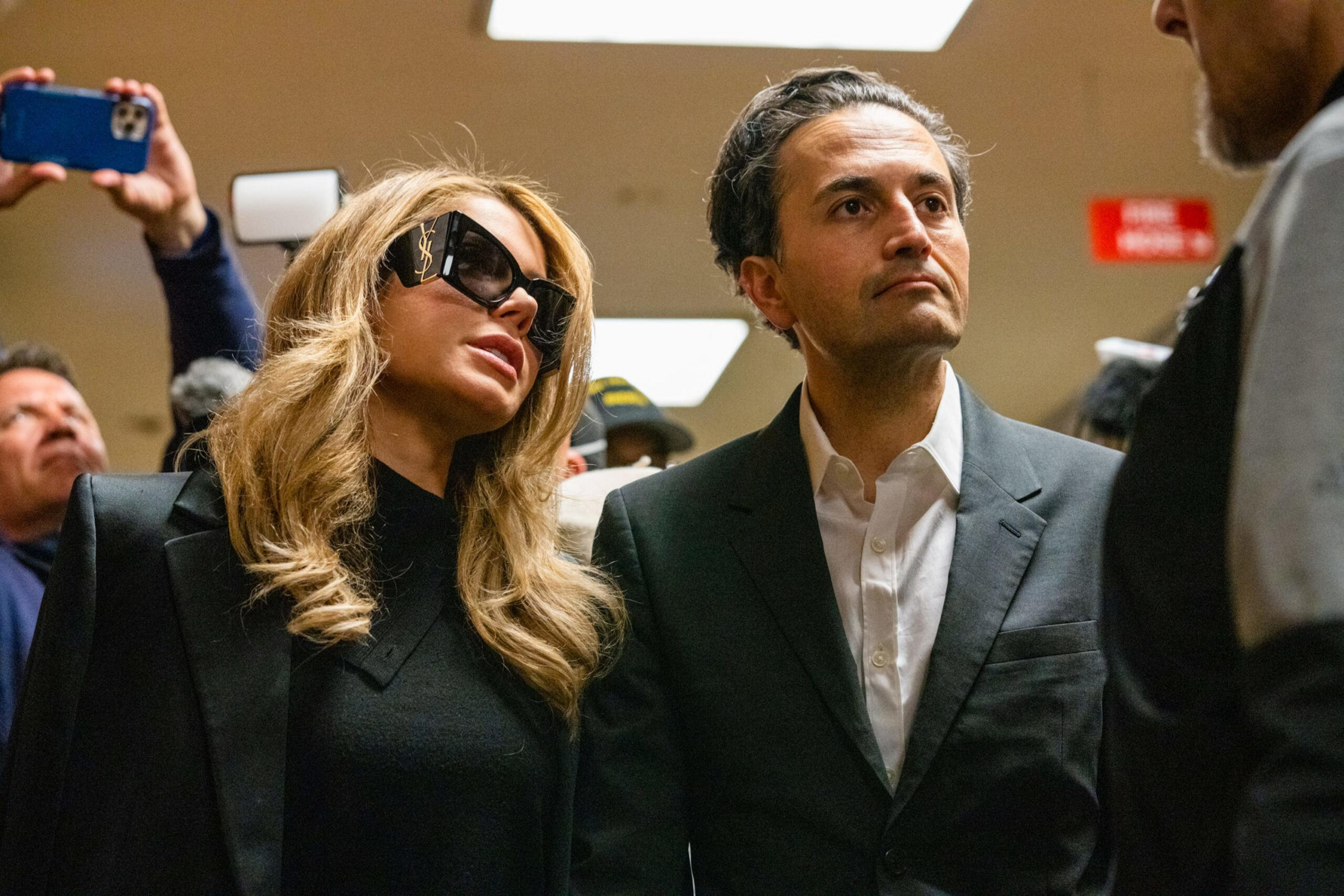 A woman with long blonde hair and large sunglasses stands next to a man in a dark suit and white shirt in a crowded indoor space.