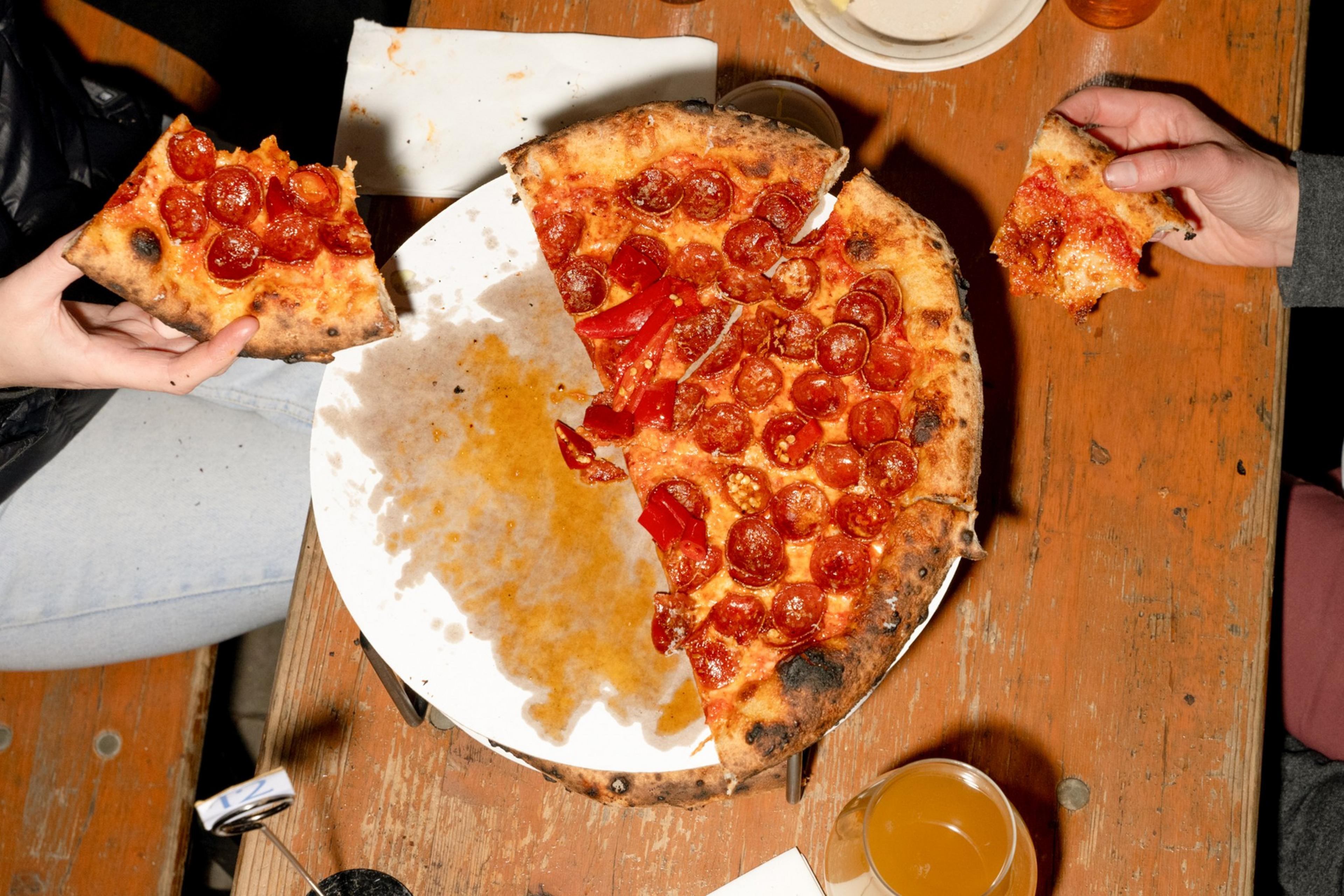 A partially eaten pepperoni pizza rests on a white plate on a wooden table, with two people each holding a slice, and a drink nearby.