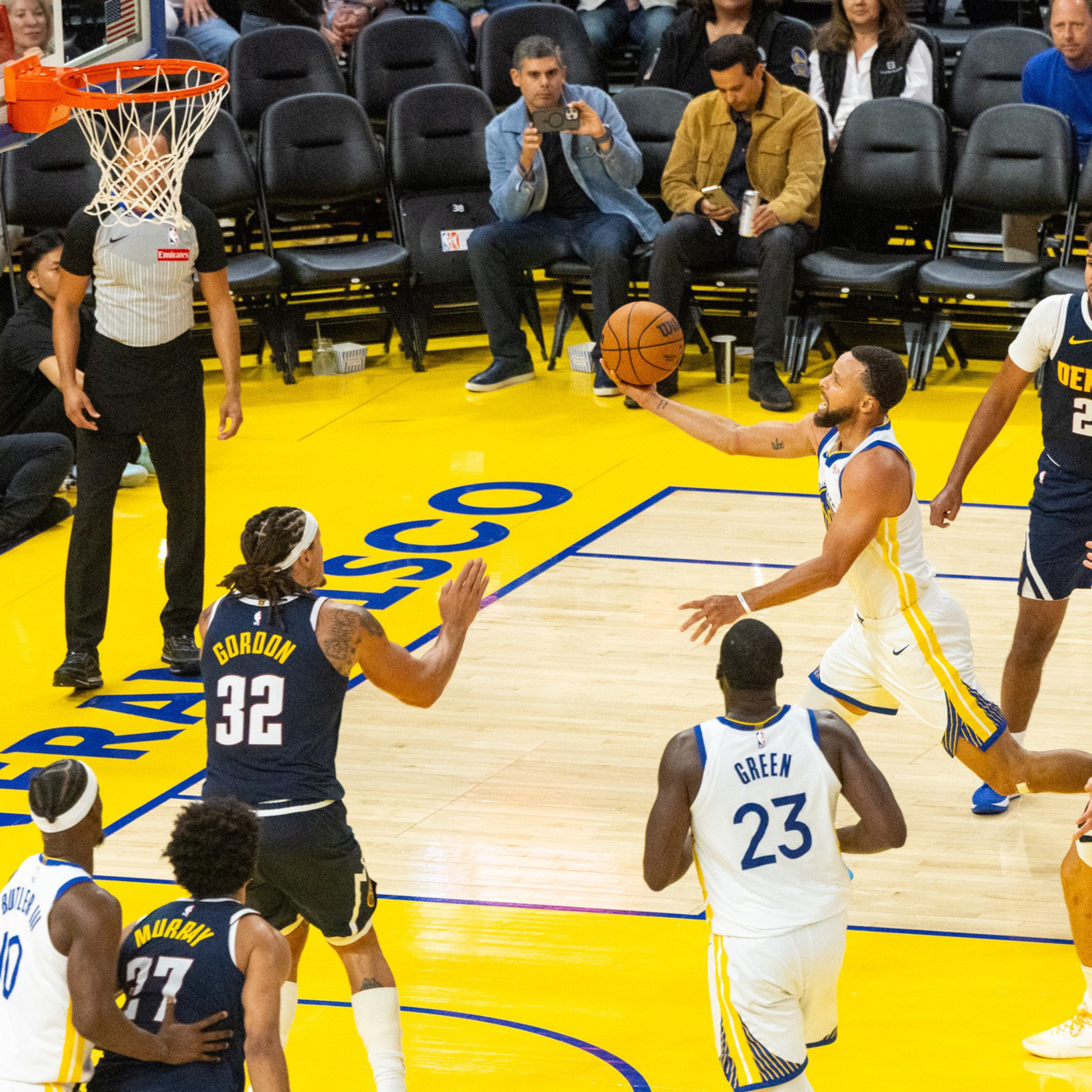 A basketball player in white and blue leaps to shoot near the hoop while four opposing players in navy and yellow watch on a yellow court.