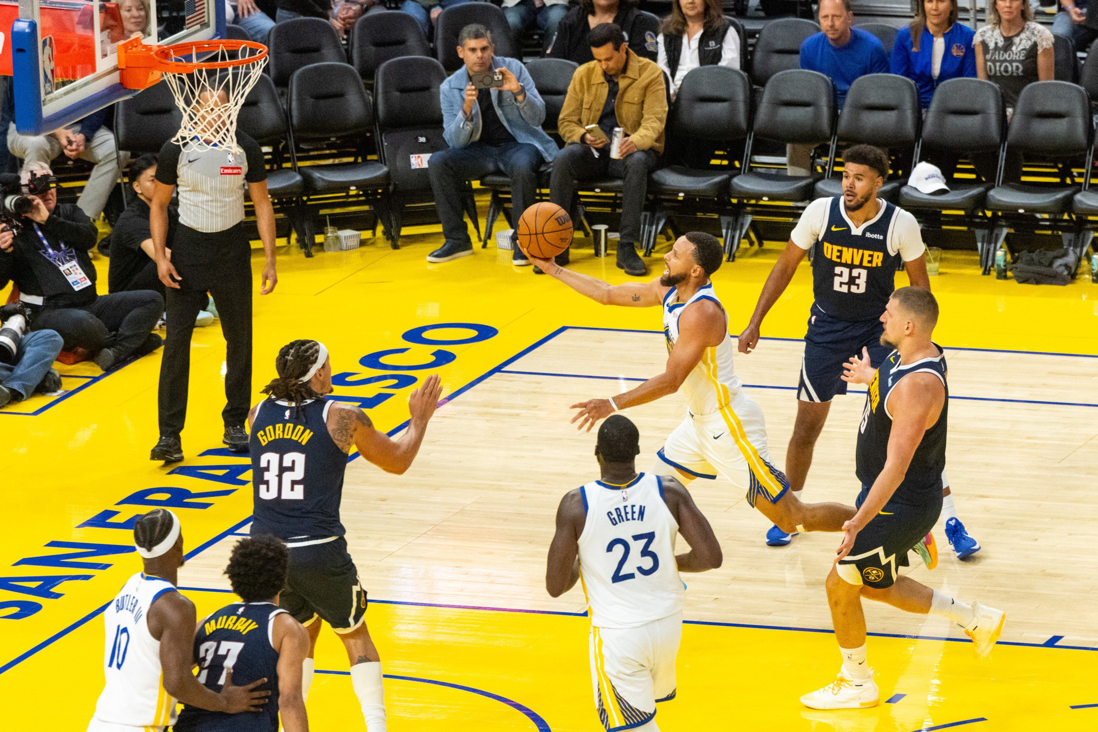 A basketball player in white and blue leaps to shoot near the hoop while four opposing players in navy and yellow watch on a yellow court.