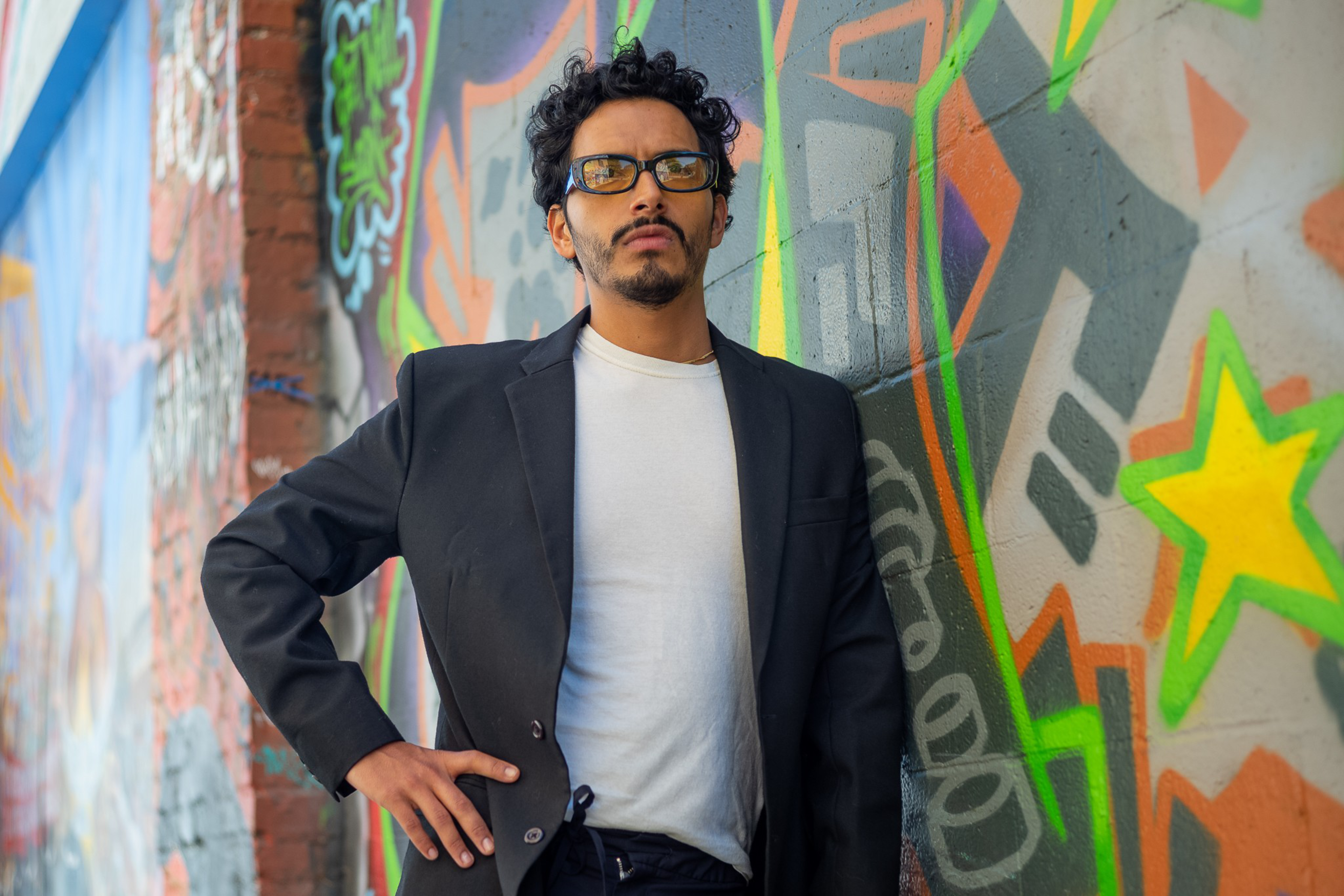 A man in a black blazer and white shirt stands confidently in front of a colorful graffiti-covered wall, with vibrant shapes and stars in the background.