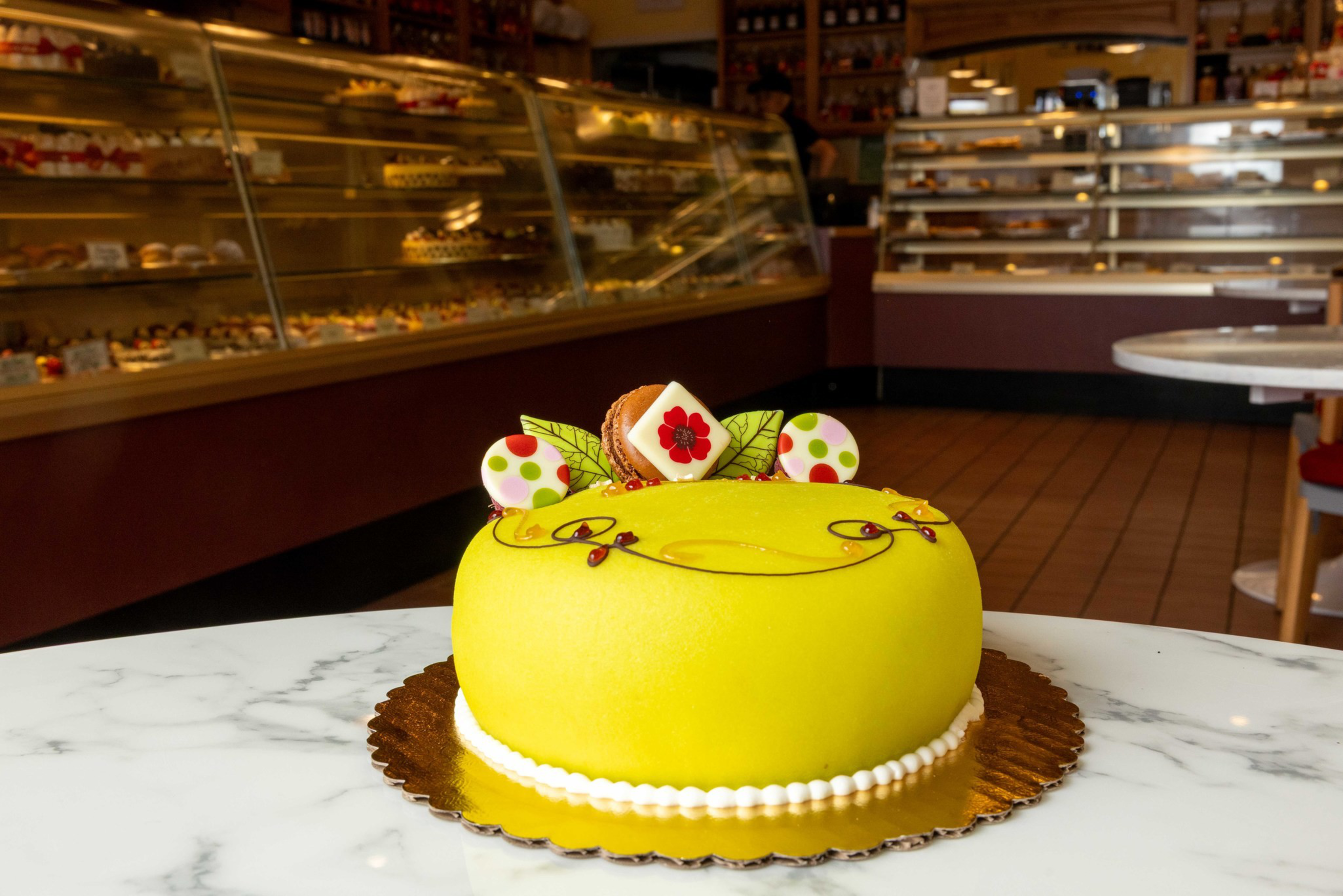 A round yellow cake with decorative polka-dot and floral toppers sits on a gold cake board atop a white marble table in a bakery.