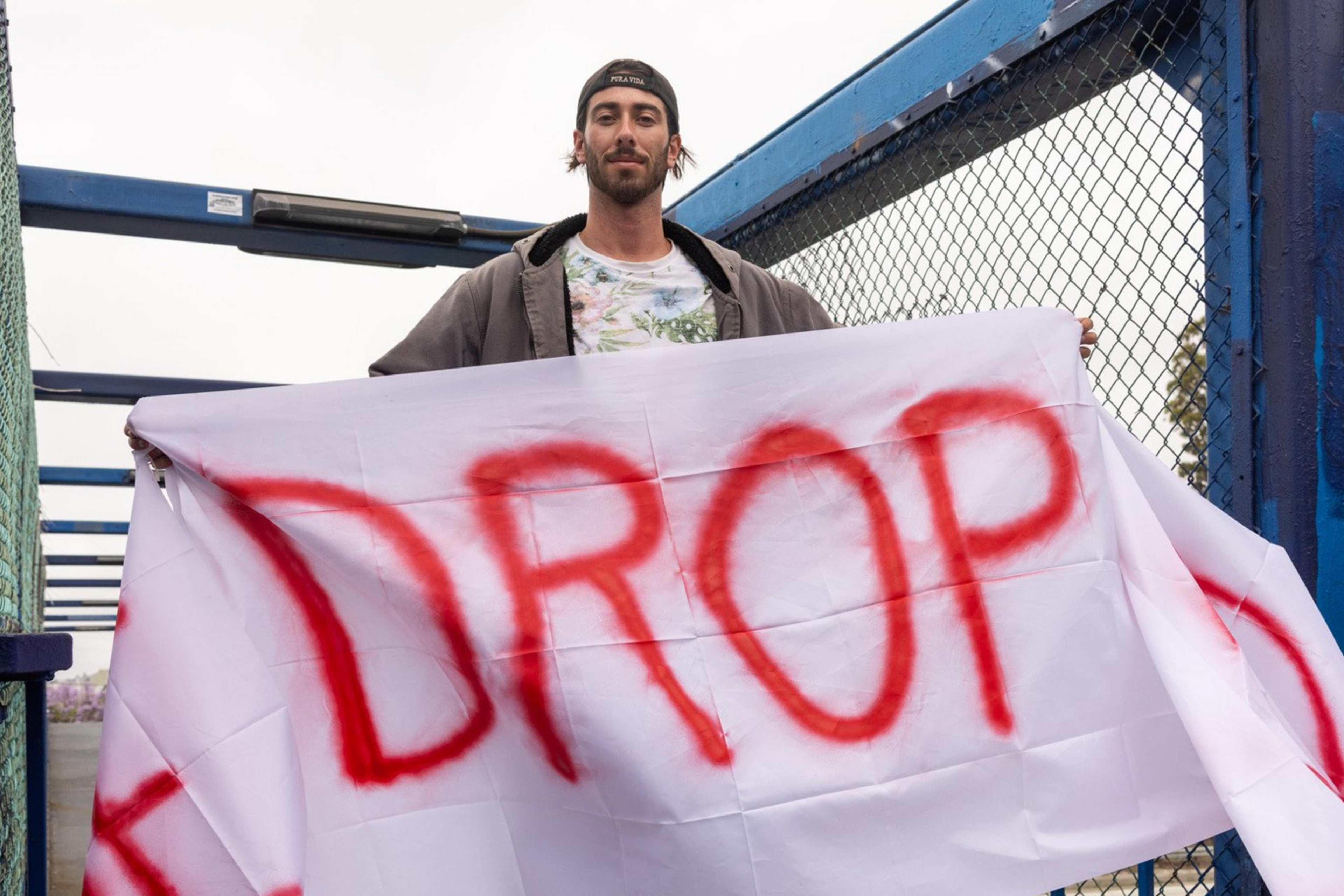 A person in a backward cap holds a large white banner with the word "DROP" spray-painted in red. They are standing on a bridge with blue fencing.