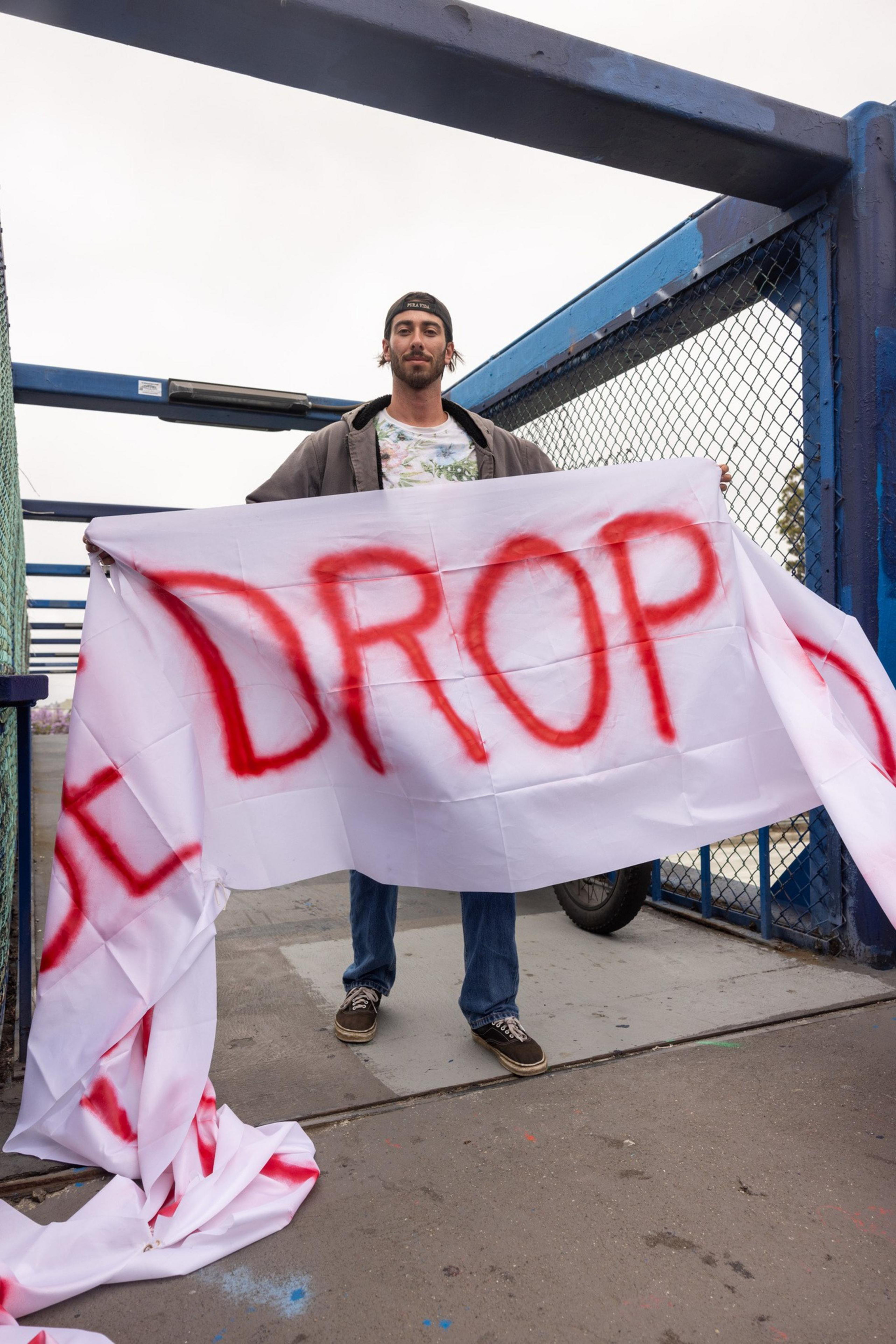 A person in a backward cap holds a large white banner with the word "DROP" spray-painted in red. They are standing on a bridge with blue fencing.