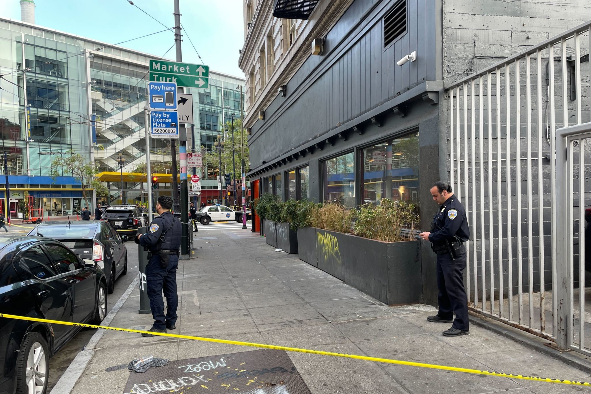 Two police officers stand on a cordoned-off urban sidewalk. One officer faces a parking sign while the other checks a device. Buildings and a parking area are visible.