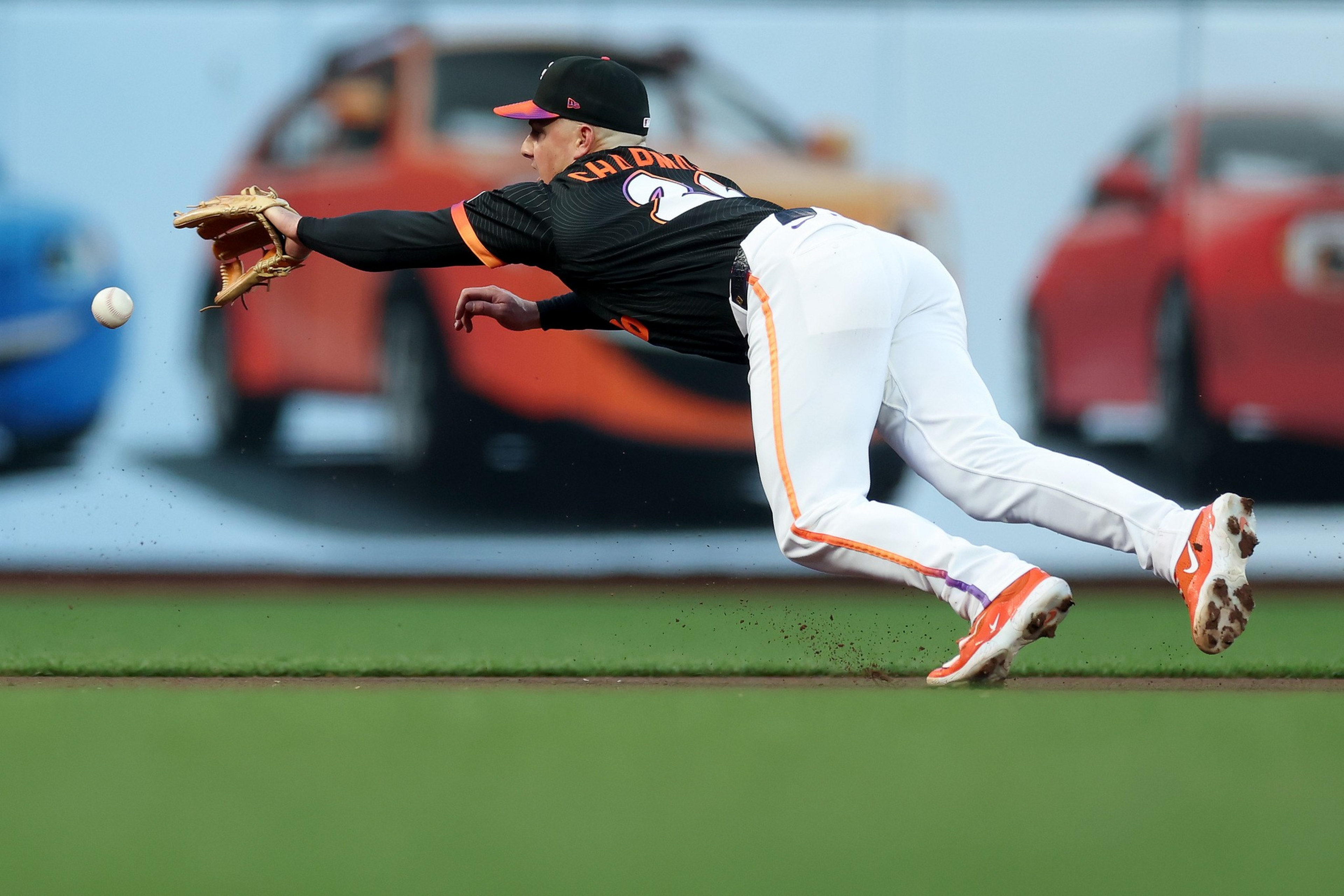 A baseball player in mid-air dive reaches with his glove to catch a ball, wearing a black and orange uniform with white pants.