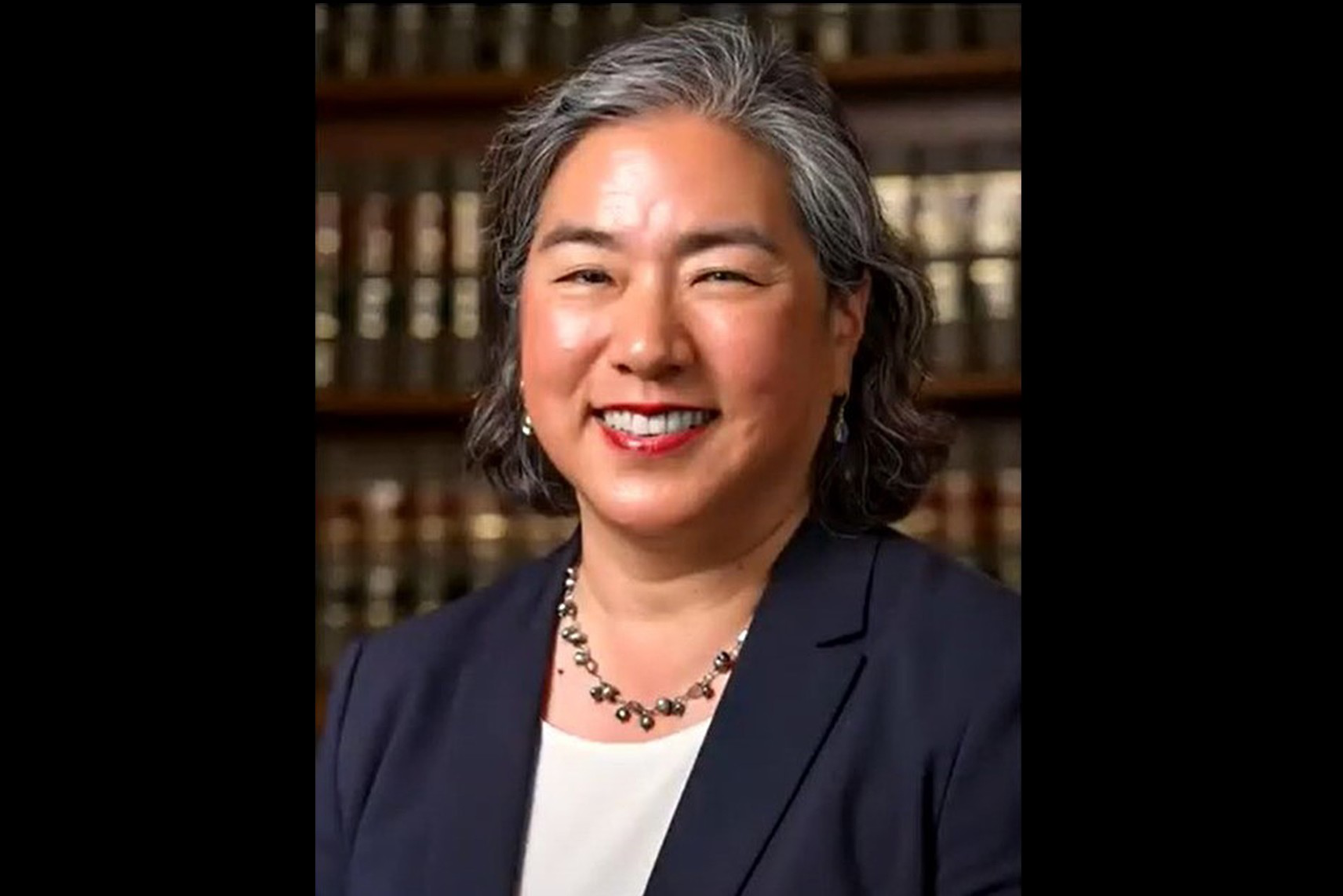 A middle-aged woman with gray-streaked hair smiles, wearing a dark blazer, white top, and necklace, standing in front of a bookshelf filled with books.