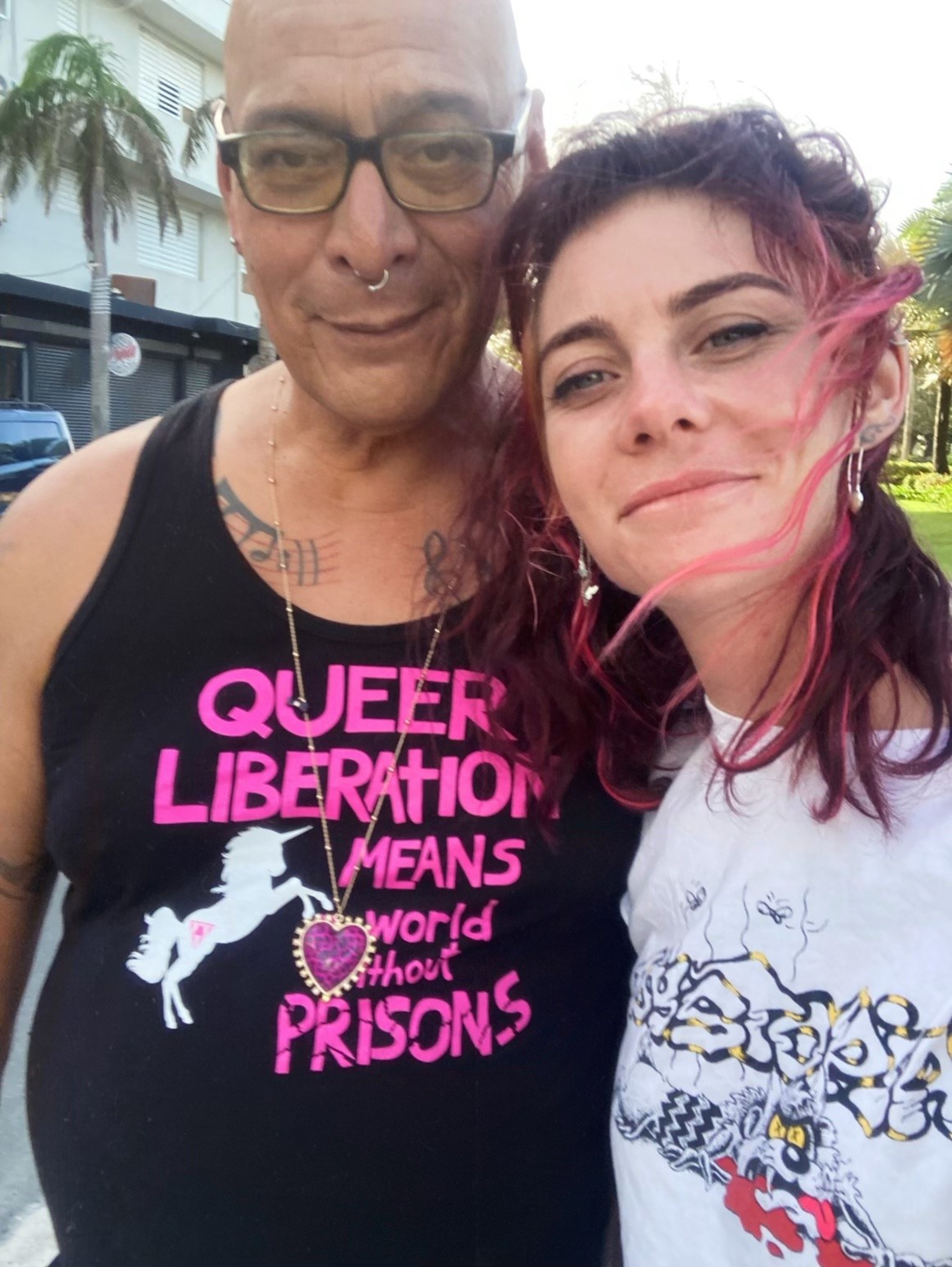A person with glasses and a nose ring wears a black tank top with pink text, standing next to a smiling person with pink-streaked hair wearing a white shirt.