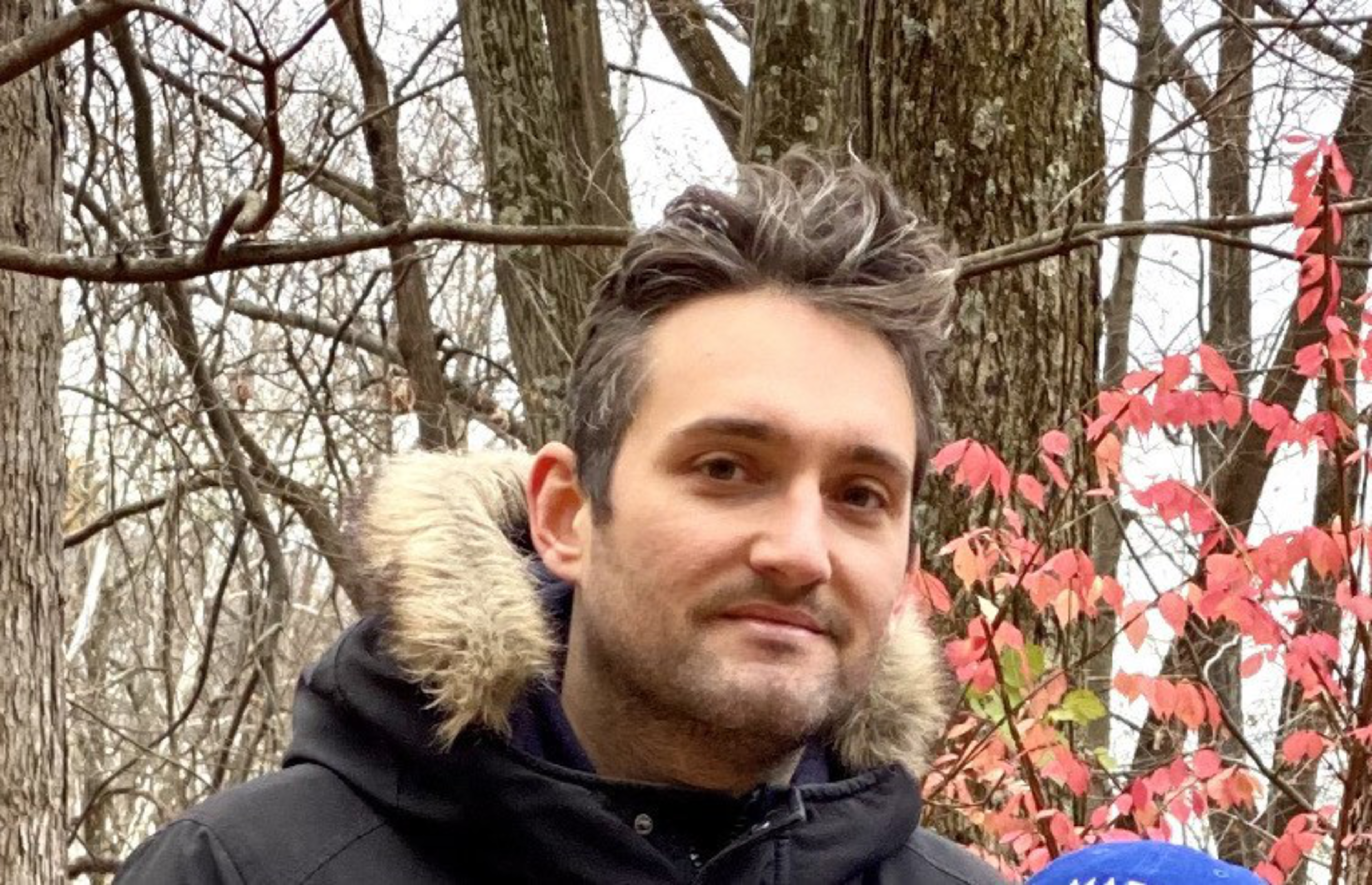 A man with short dark hair and light facial hair wears a black coat with fur trim outdoors, surrounded by bare tree branches and red leaves.