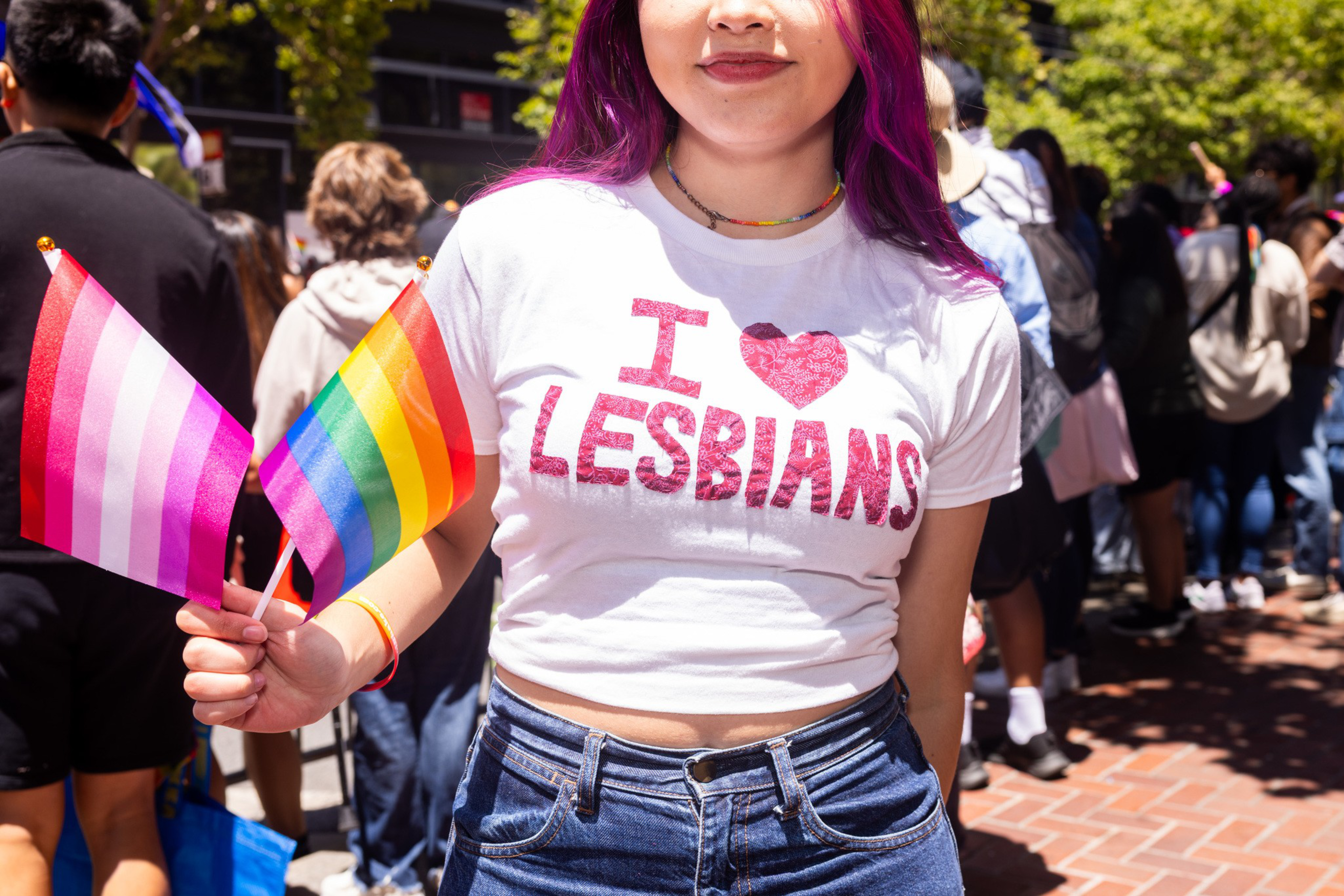 A person with purple hair wears a white shirt saying &quot;I ♥ LESBIANS&quot; and holds lesbian and rainbow flags, standing in a crowded outdoor setting.