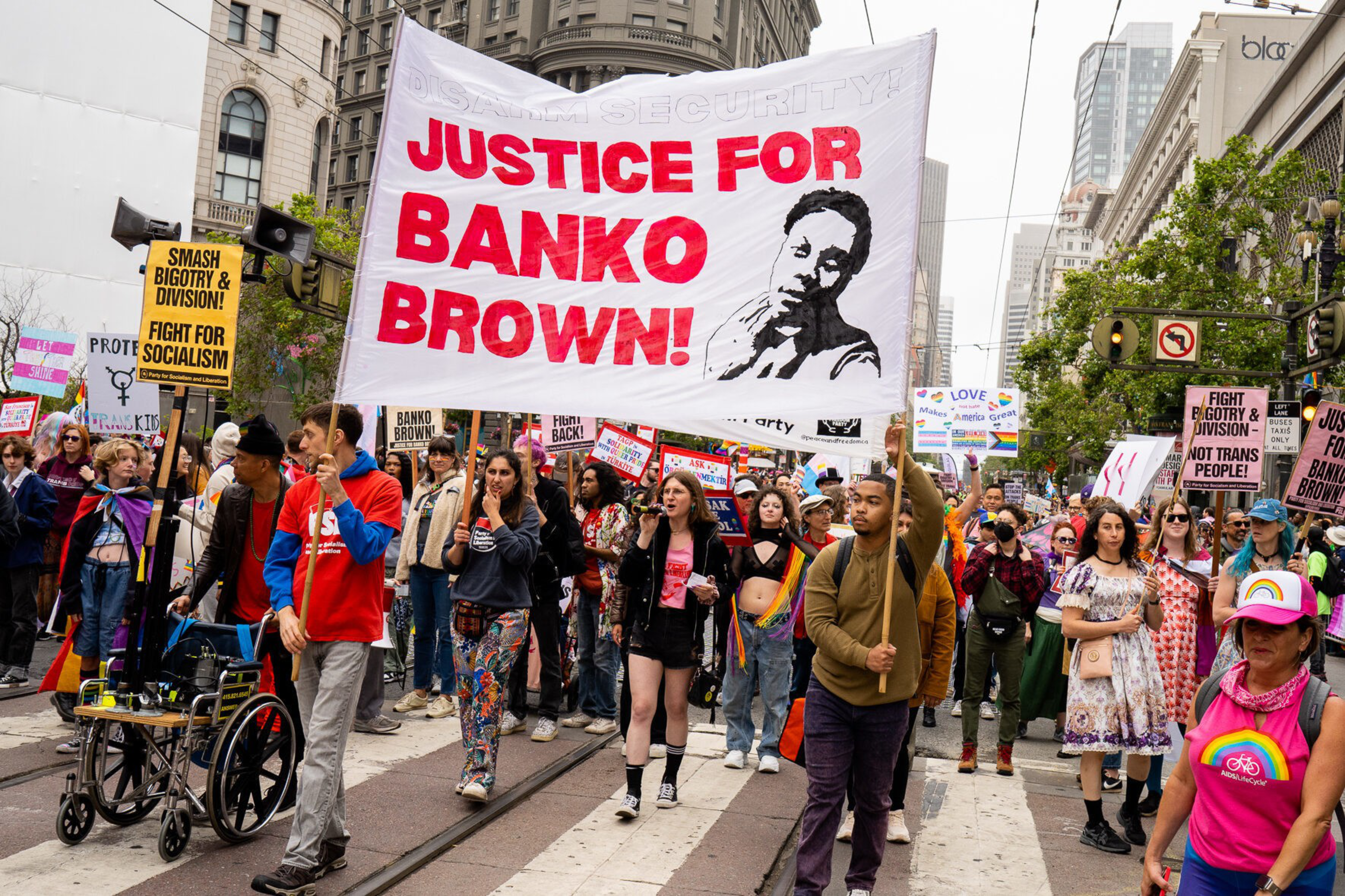 A diverse crowd marches with signs advocating justice for Banko Brown and fighting bigotry, socialism, and division in an urban street setting.