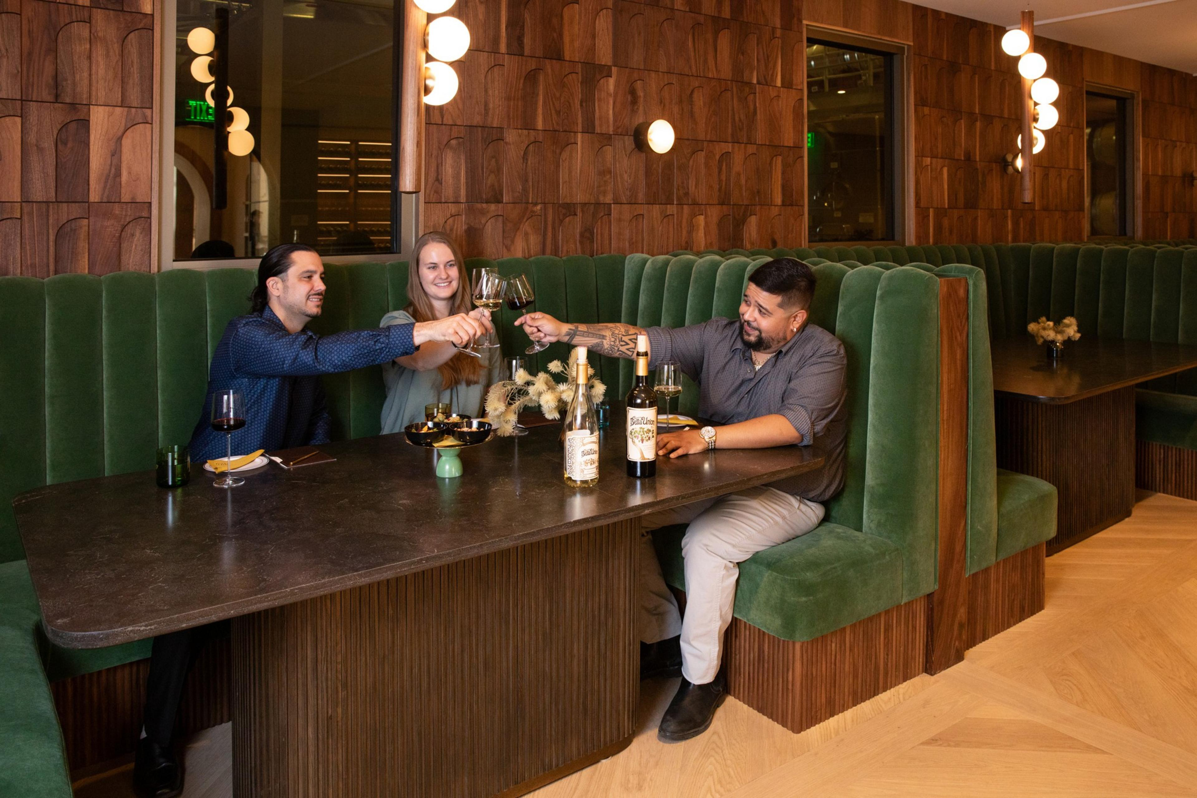 Three people sit in a cozy booth, clinking wine glasses. The setting has green velvet seats and wooden decor, with bottles and snacks on the table.