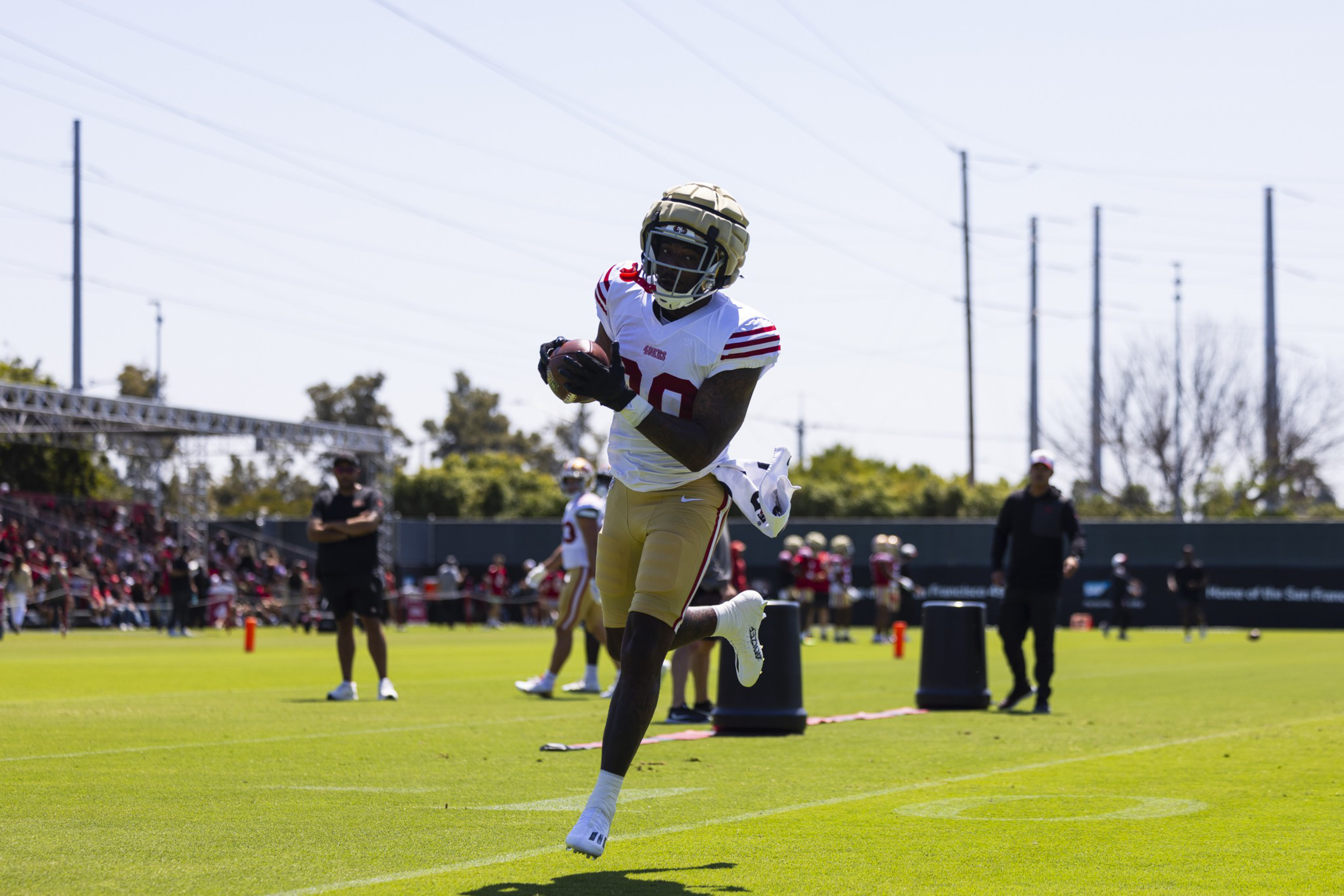 A football player wearing a gold helmet and white jersey catches a ball mid-air on a sunny field with teammates and coaches in the background.