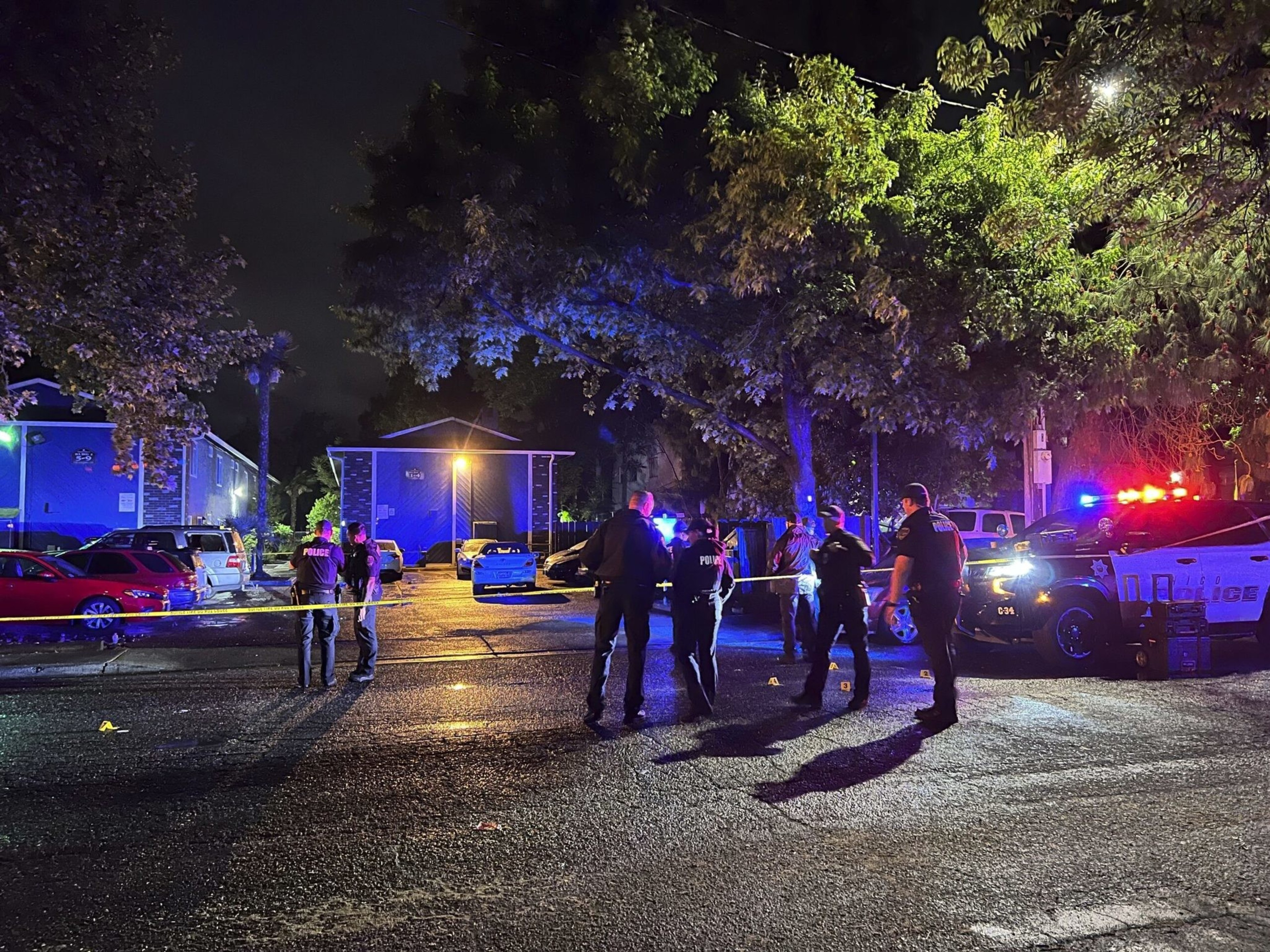 Police officers stand behind yellow caution tape in a dimly lit parking lot at night, with police vehicles flashing red and blue lights nearby.