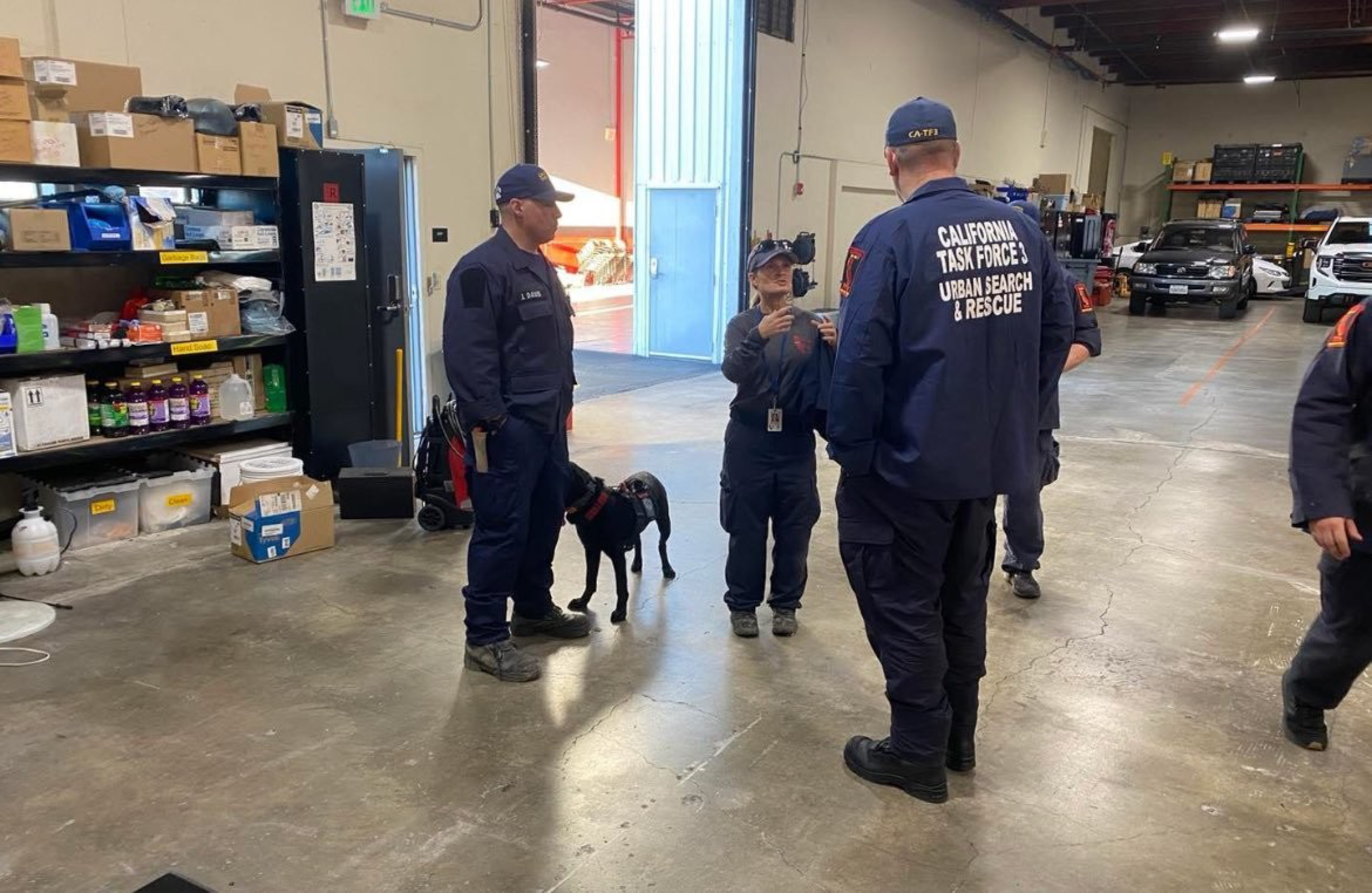 Four people in navy uniforms, labeled &quot;California Task Force 3 Urban Search &amp; Rescue,&quot; are in a warehouse with a black dog, surrounded by shelves and vehicles.