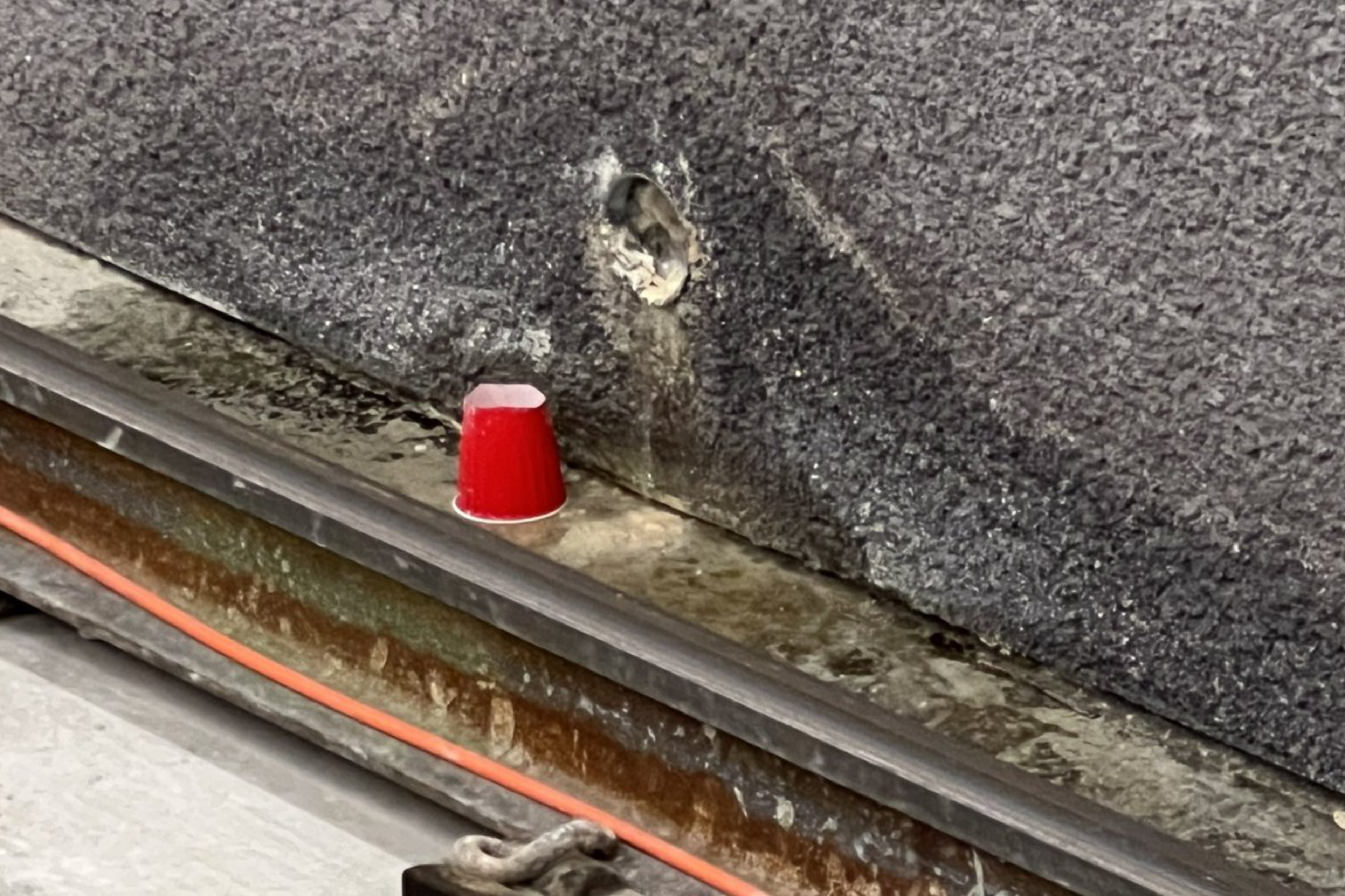 A small red plastic cup sits upside down on a ledge against a rough dark wall with a hole above it, next to rusty metal rails and an orange cable.