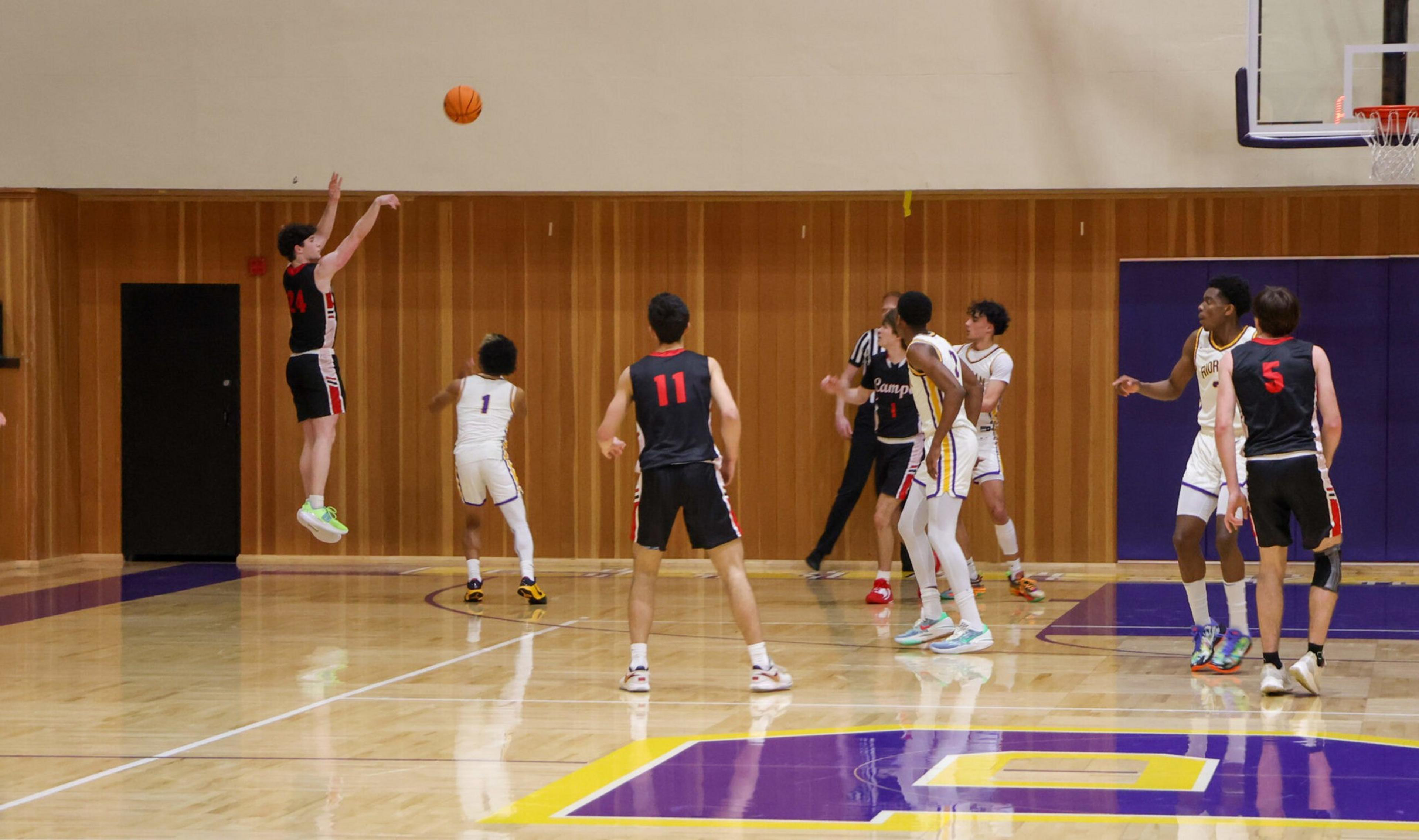 A basketball player in a black uniform jumps to shoot while players in white and black uniforms watch near the basket on a polished indoor court.