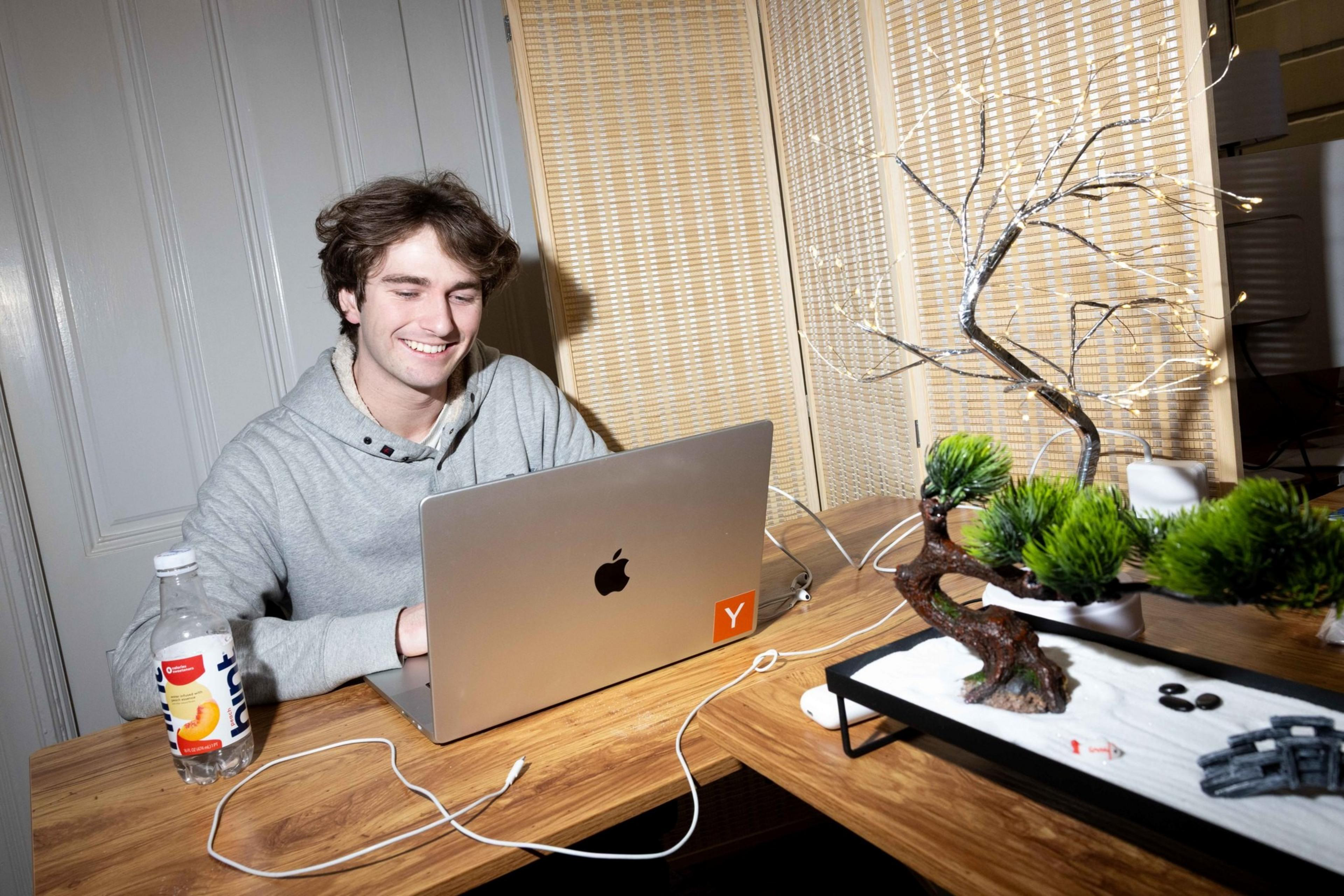 A person in a gray hoodie smiles while using a laptop on a wooden table. Nearby, there’s a bottle of water and a decorative bonsai tree with lights.