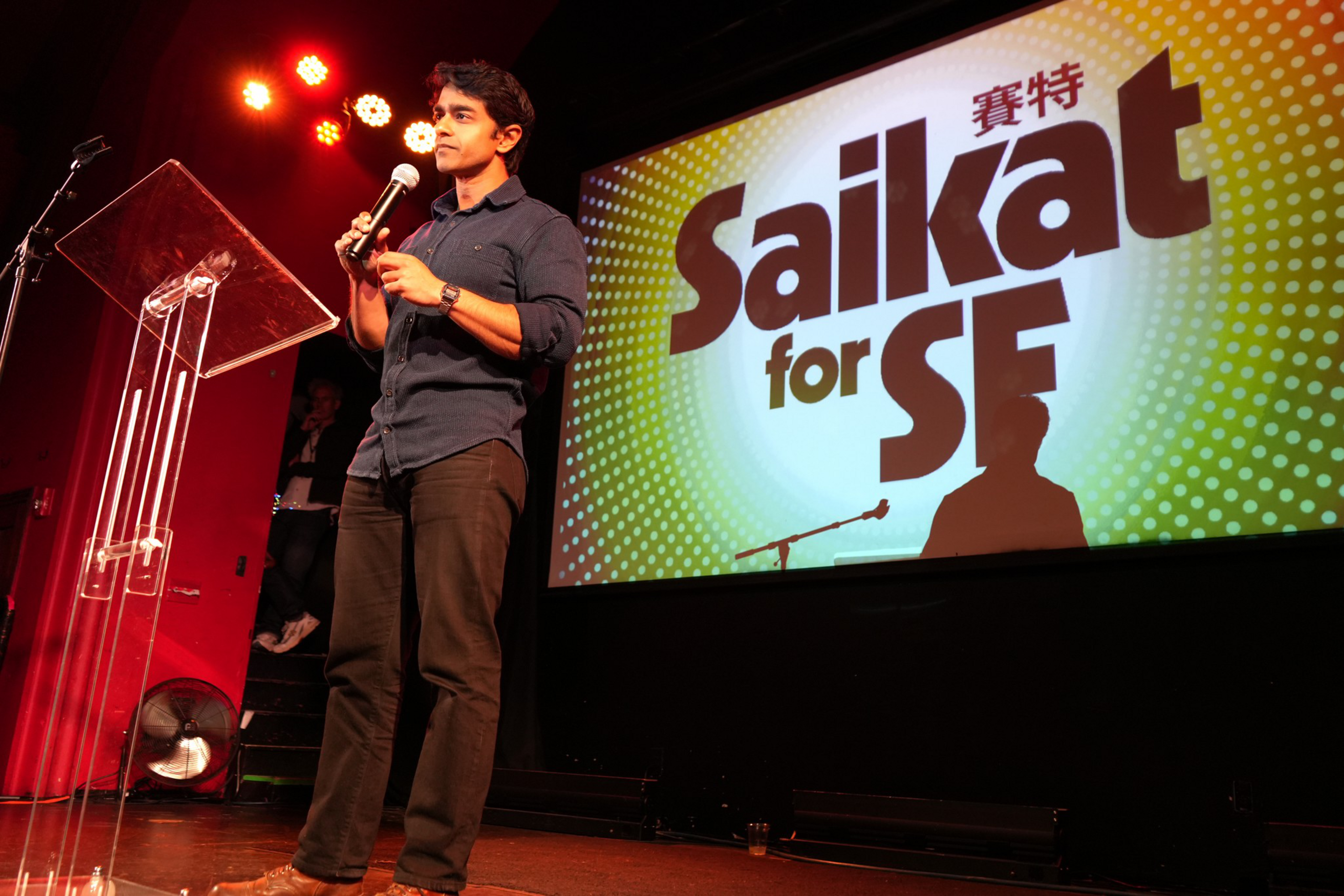 A man stands on stage holding a microphone near a clear podium, with a colorful screen behind him displaying the text “Saikat for SF.”