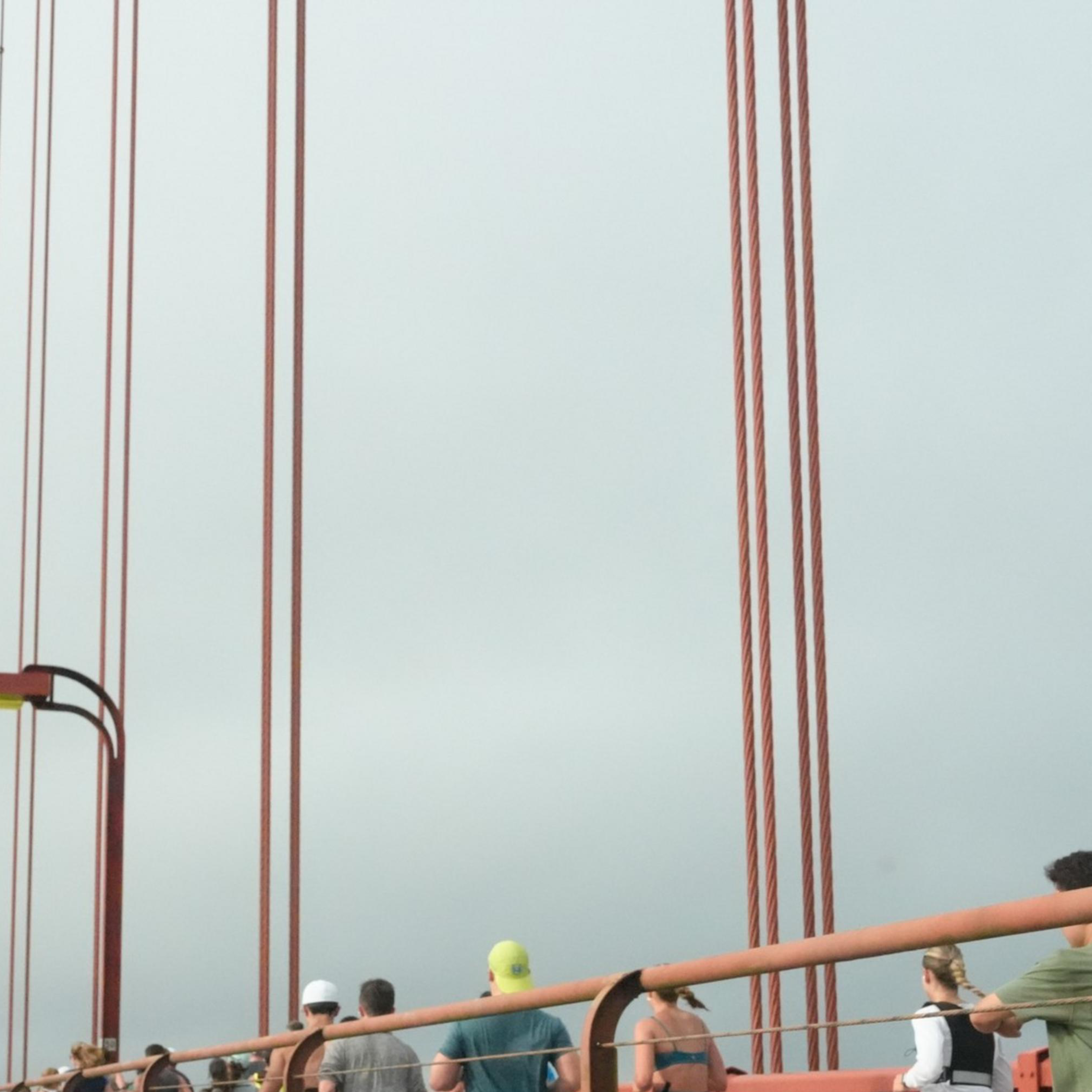 People are jogging along the pedestrian path of a large red suspension bridge on a cloudy day, while cars drive on the adjacent road.