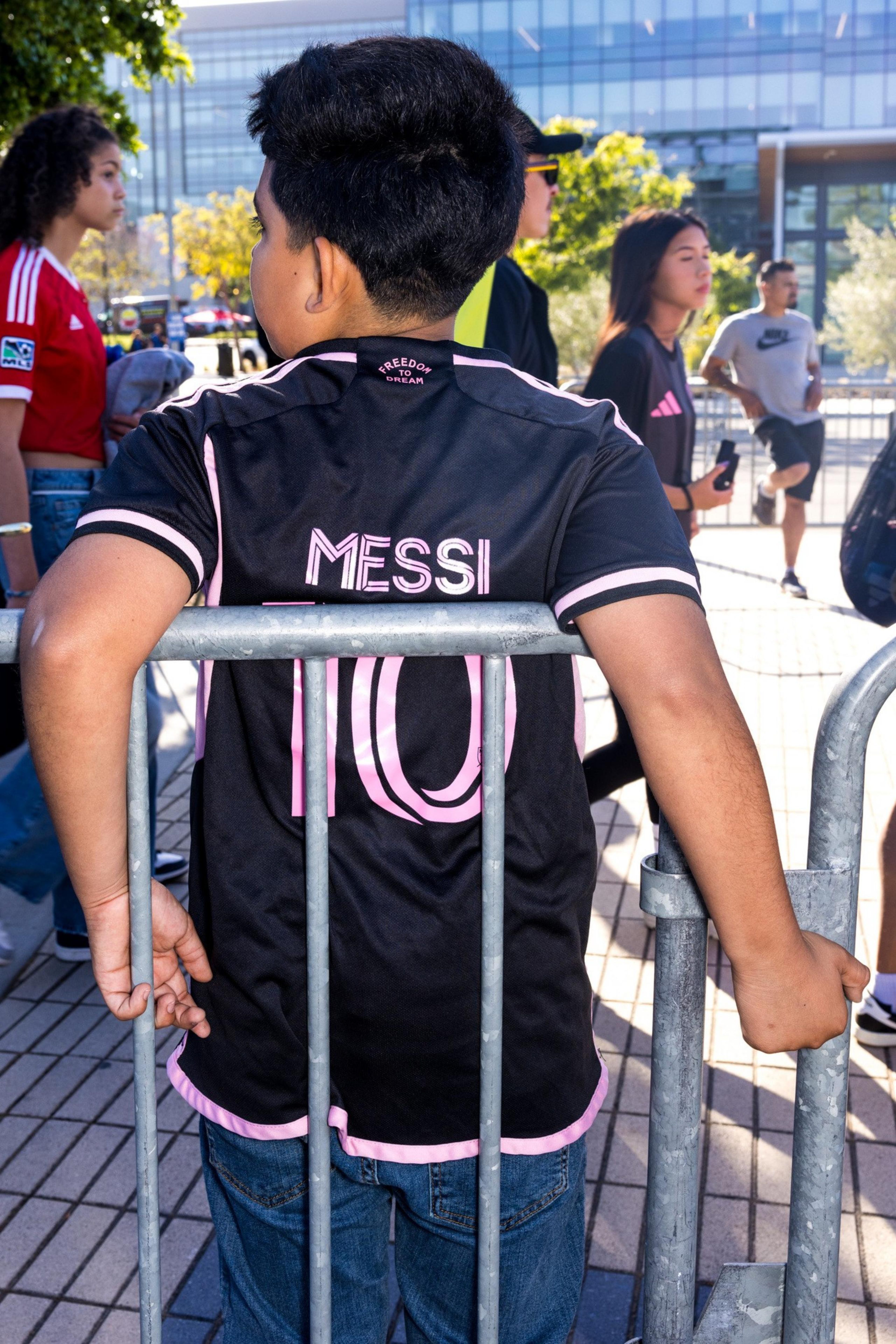 A person wearing a black and pink &quot;Messi&quot; jersey stands behind a railing, looking at a group of people in casual clothing outside a modern building.