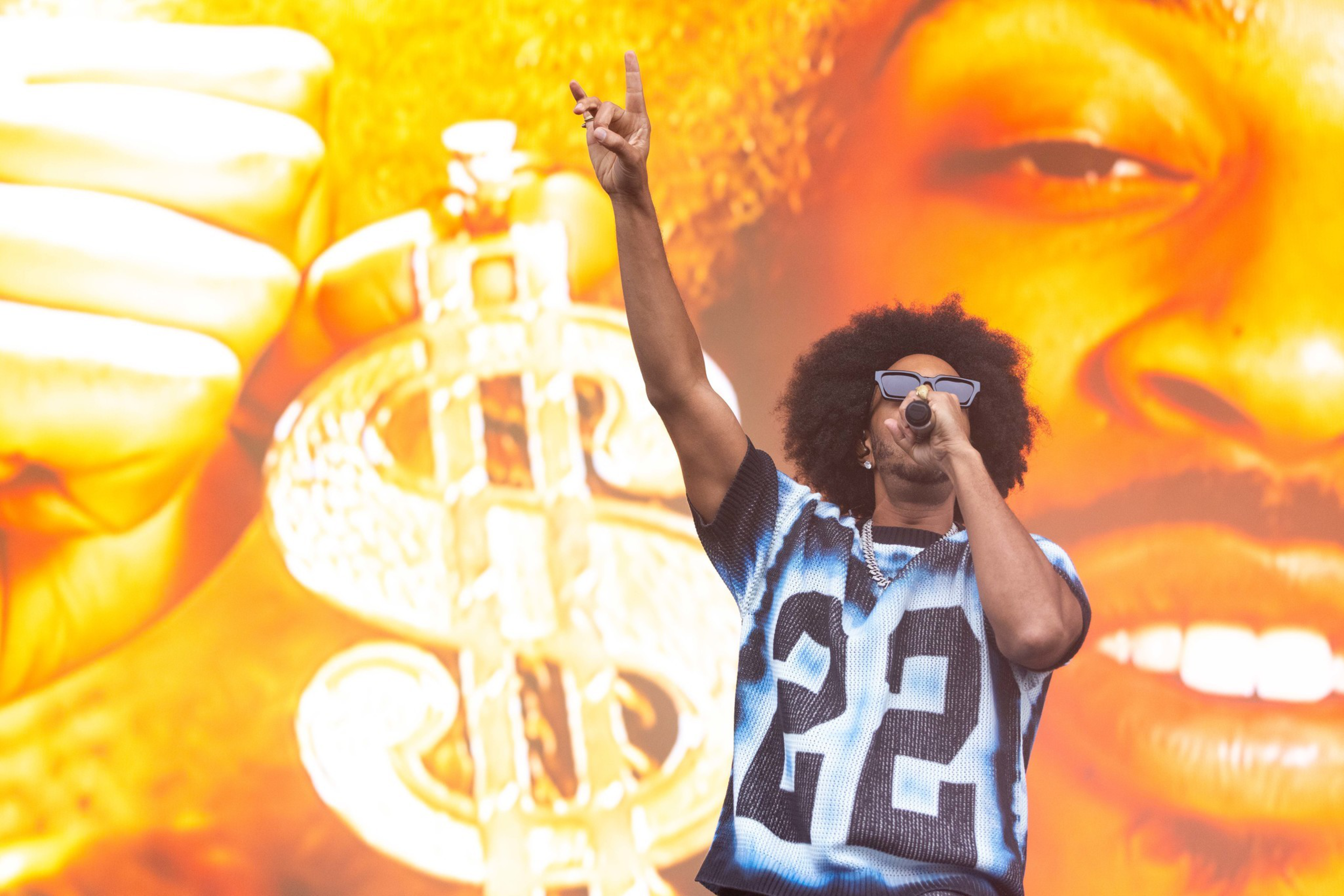 A man with an afro wearing sunglasses and a numbered jersey holds a microphone and raises his hand, standing in front of a large dollar sign backdrop.