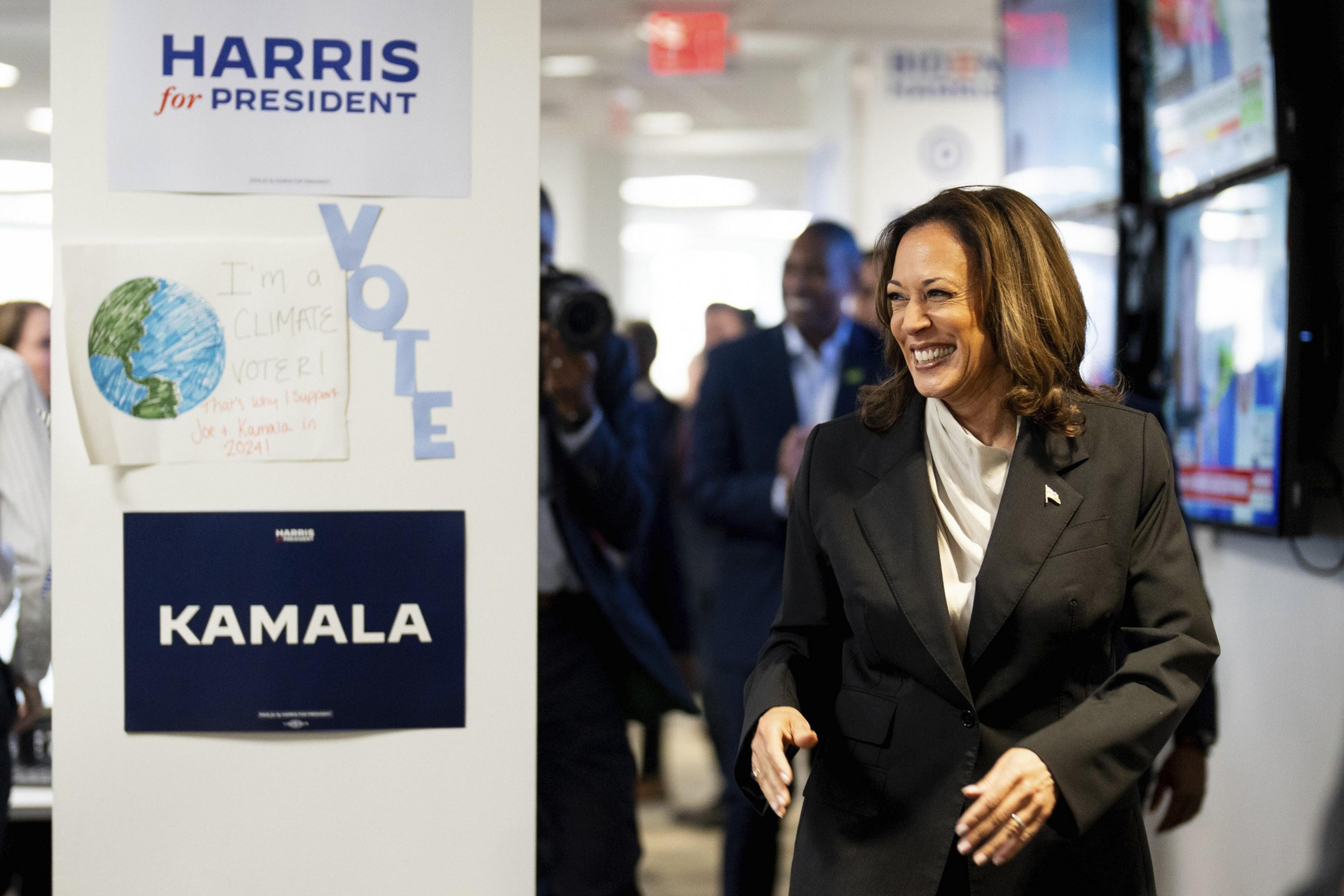 A smiling woman in a black suit stands near a wall with campaign signs for &quot;Harris for President&quot; and &quot;Kamala.&quot; Other people are in the background, some taking photos.
