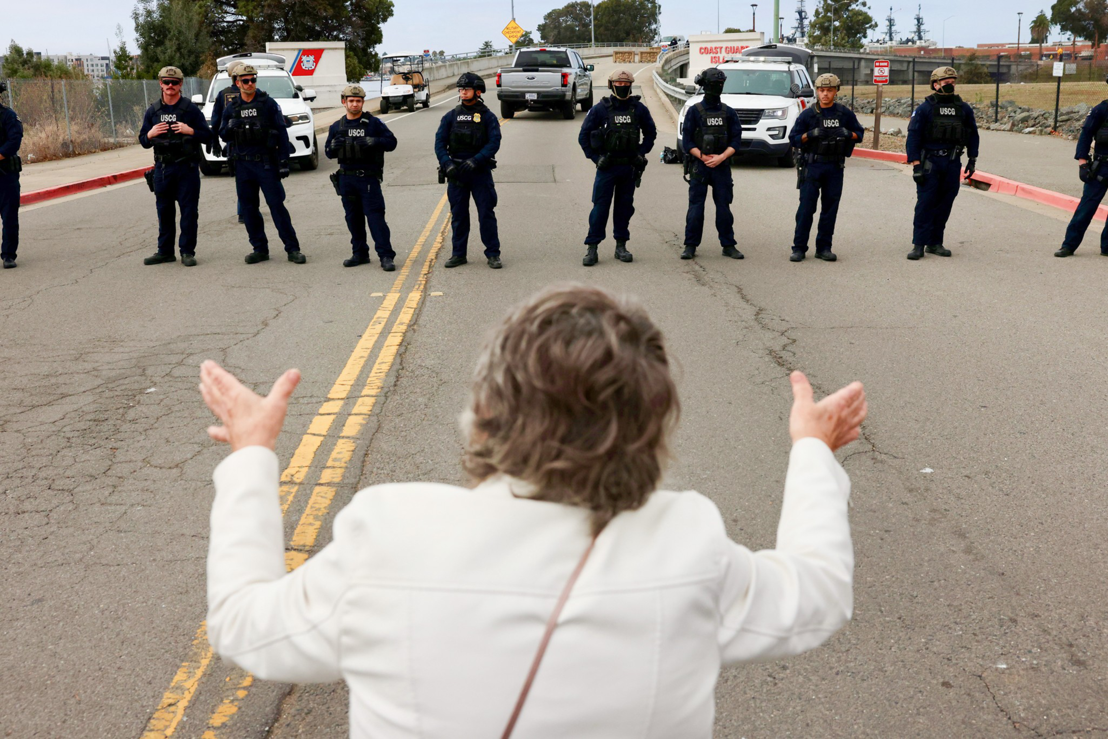 A person in a white jacket faces a line of uniformed officers blocking the road, with police vehicles and a fence visible behind them.