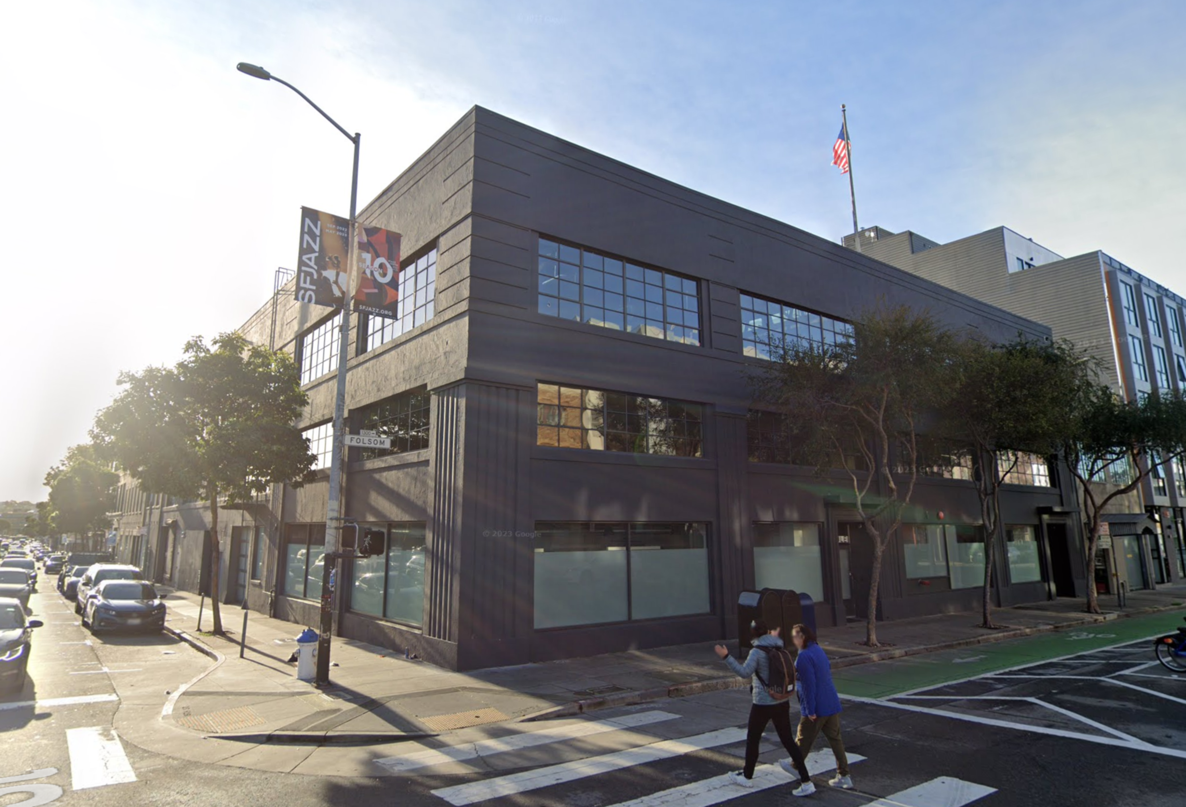 A black two-story building stands on a sunny street corner with large windows, an American flag on top, and two people crossing the crosswalk.