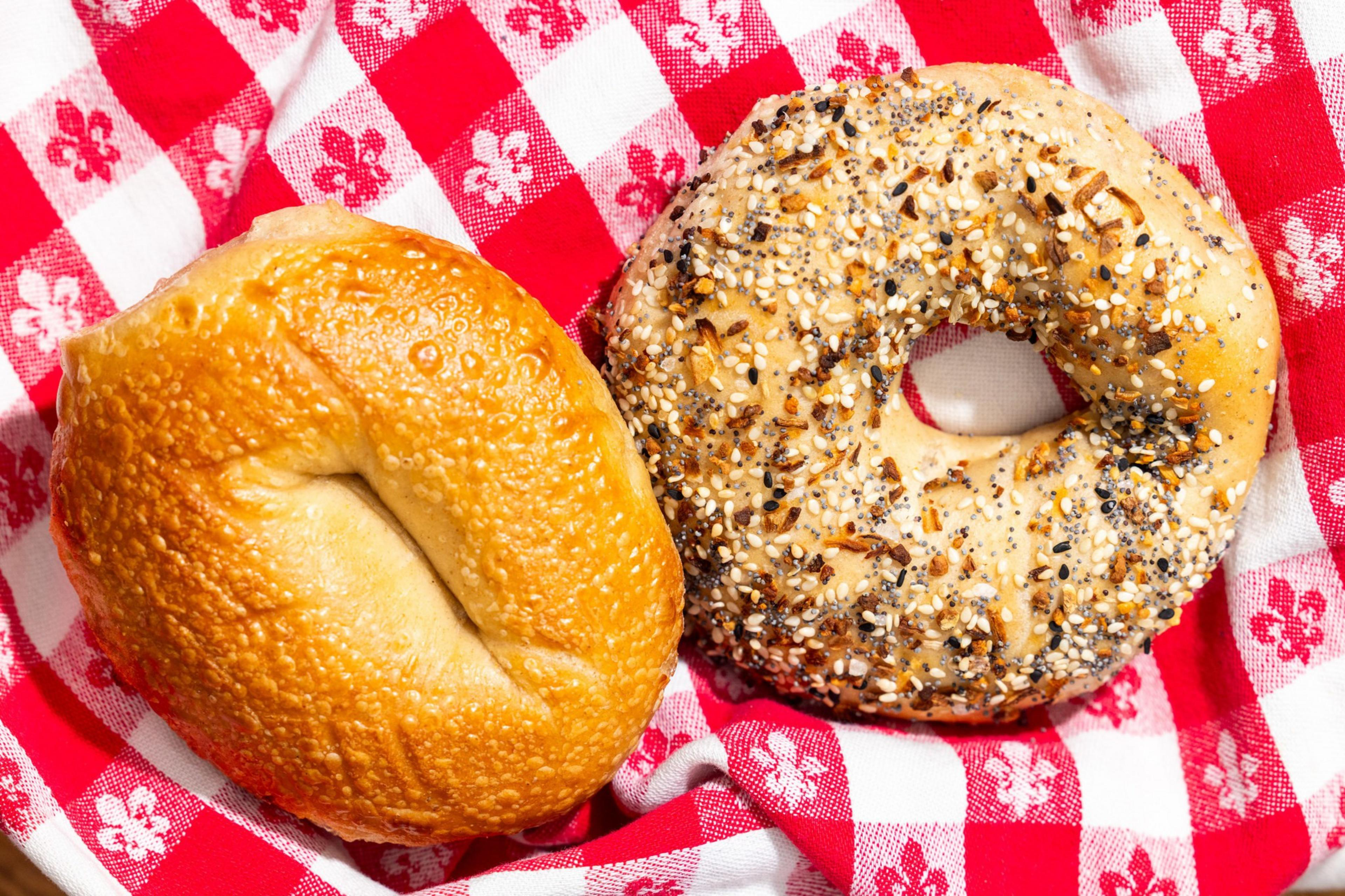 A plain bagel and a seeded bagel sit on a red and white checkered cloth with floral patterns.
