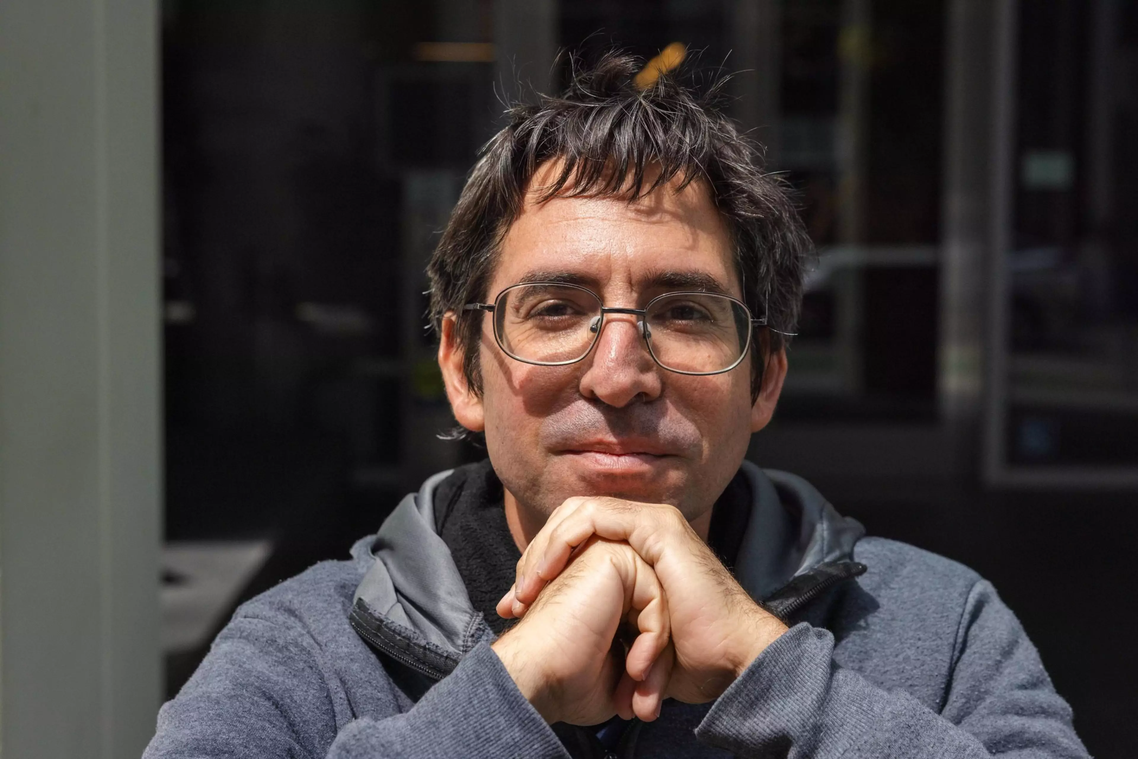 A man with glasses and tousled dark hair rests his chin on his interlaced hands, wearing a gray hoodie with a calm expression.