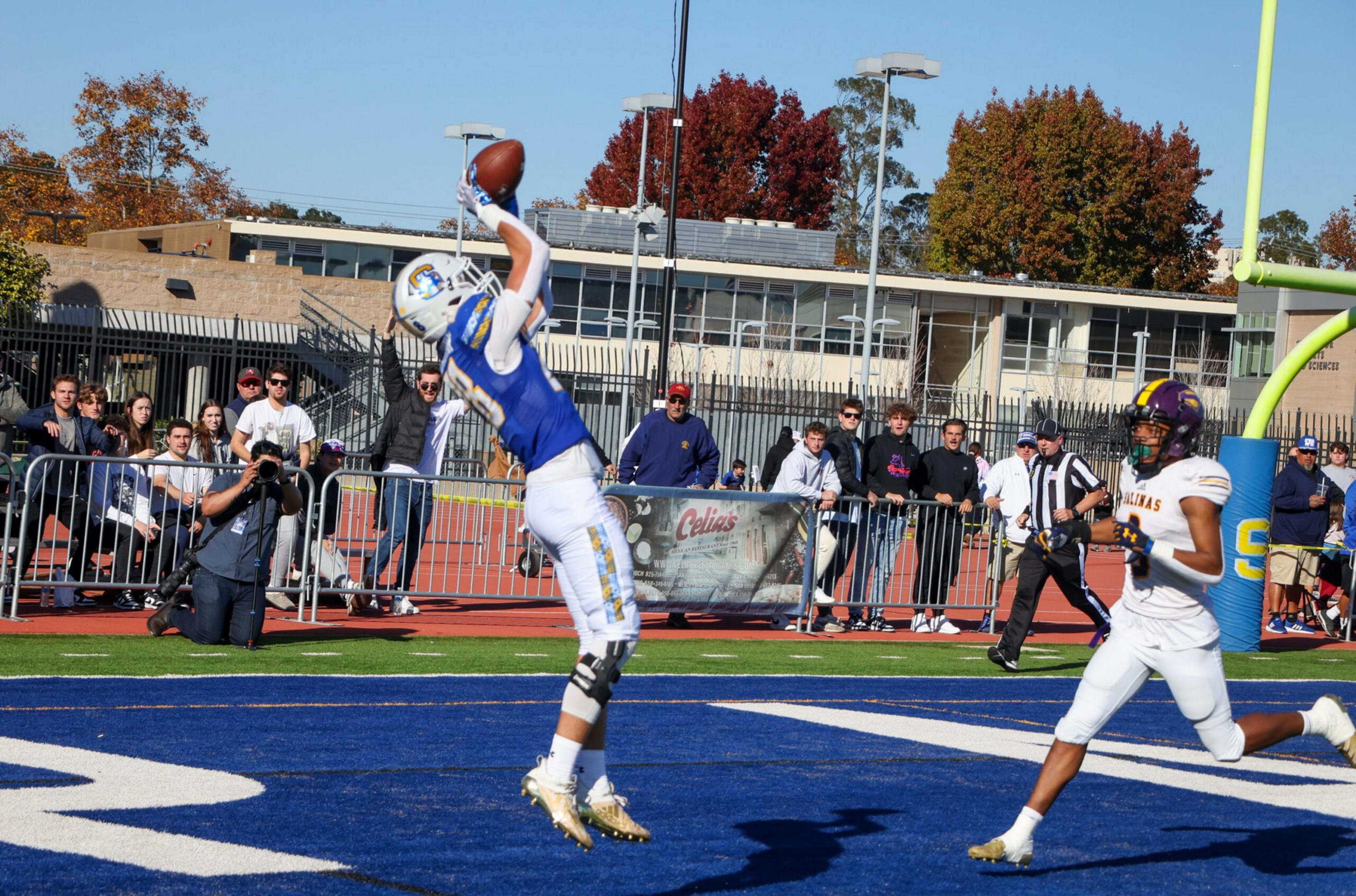 A football player in a blue and white uniform leaps to catch a pass in the end zone while a defender in white and purple runs nearby.